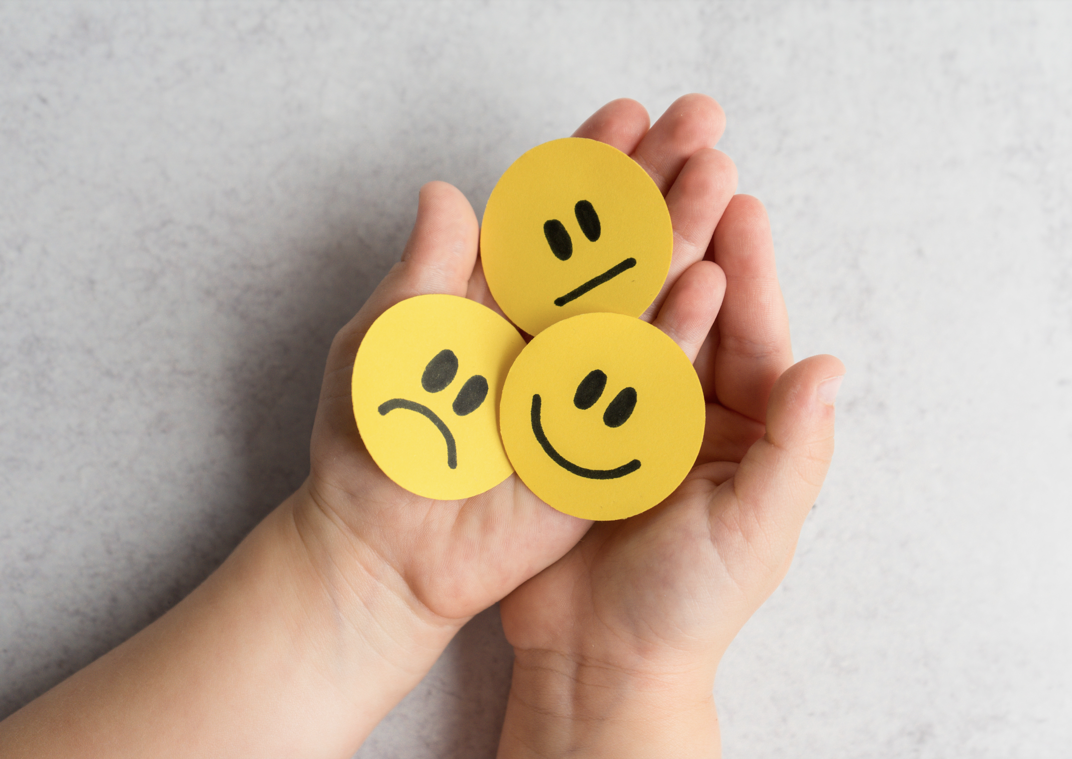 A hand holding three yellow paper circles with black emoticon faces, showing a sad face, a neutral face, and a happy face, against a light grey background.