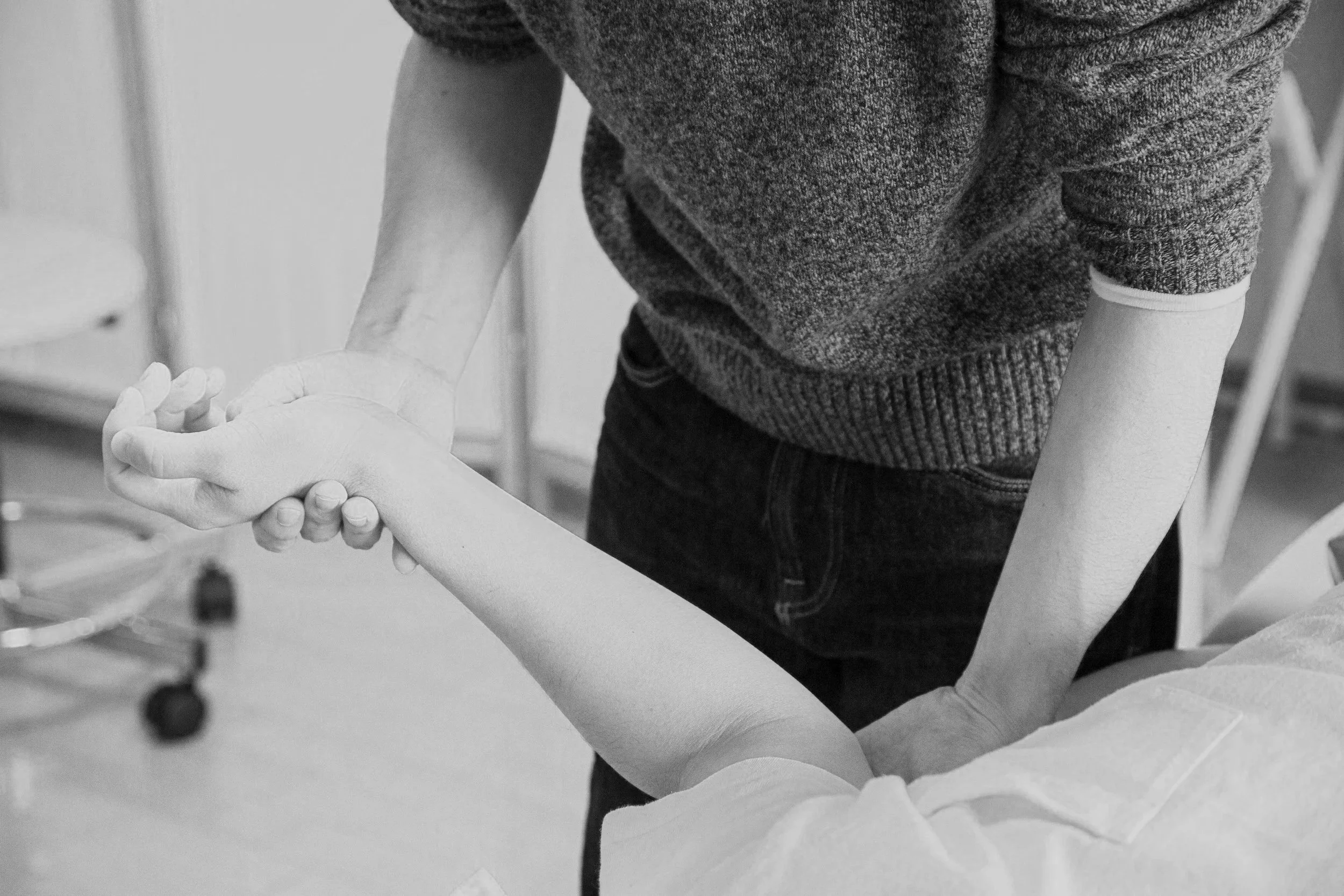 A person holding and supporting an outstretched arm of another person in a medical or physical therapy setting, with medical equipment visible in the background.