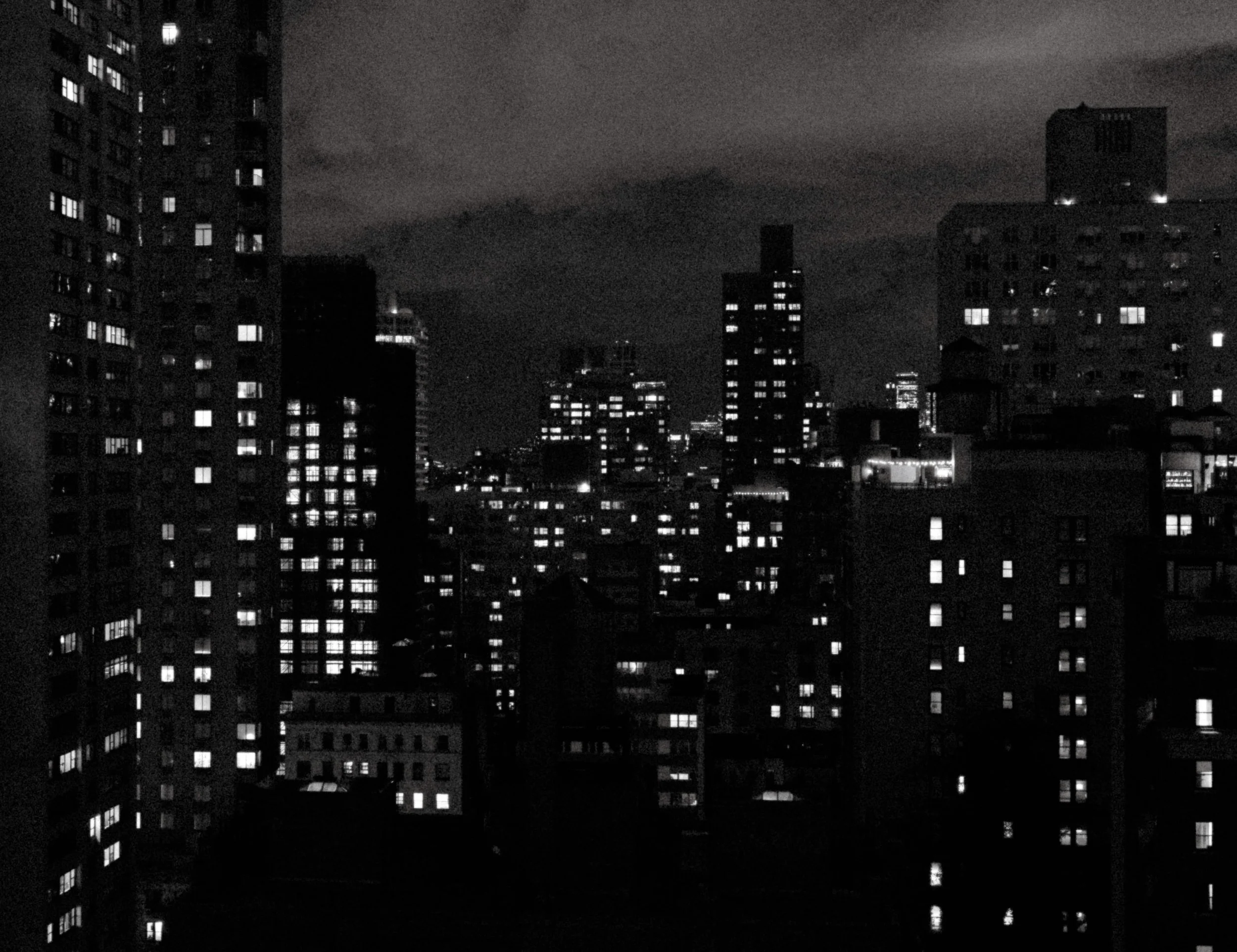Nighttime cityscape with high-rise buildings, many windows lit up, and a dark sky with clouds.