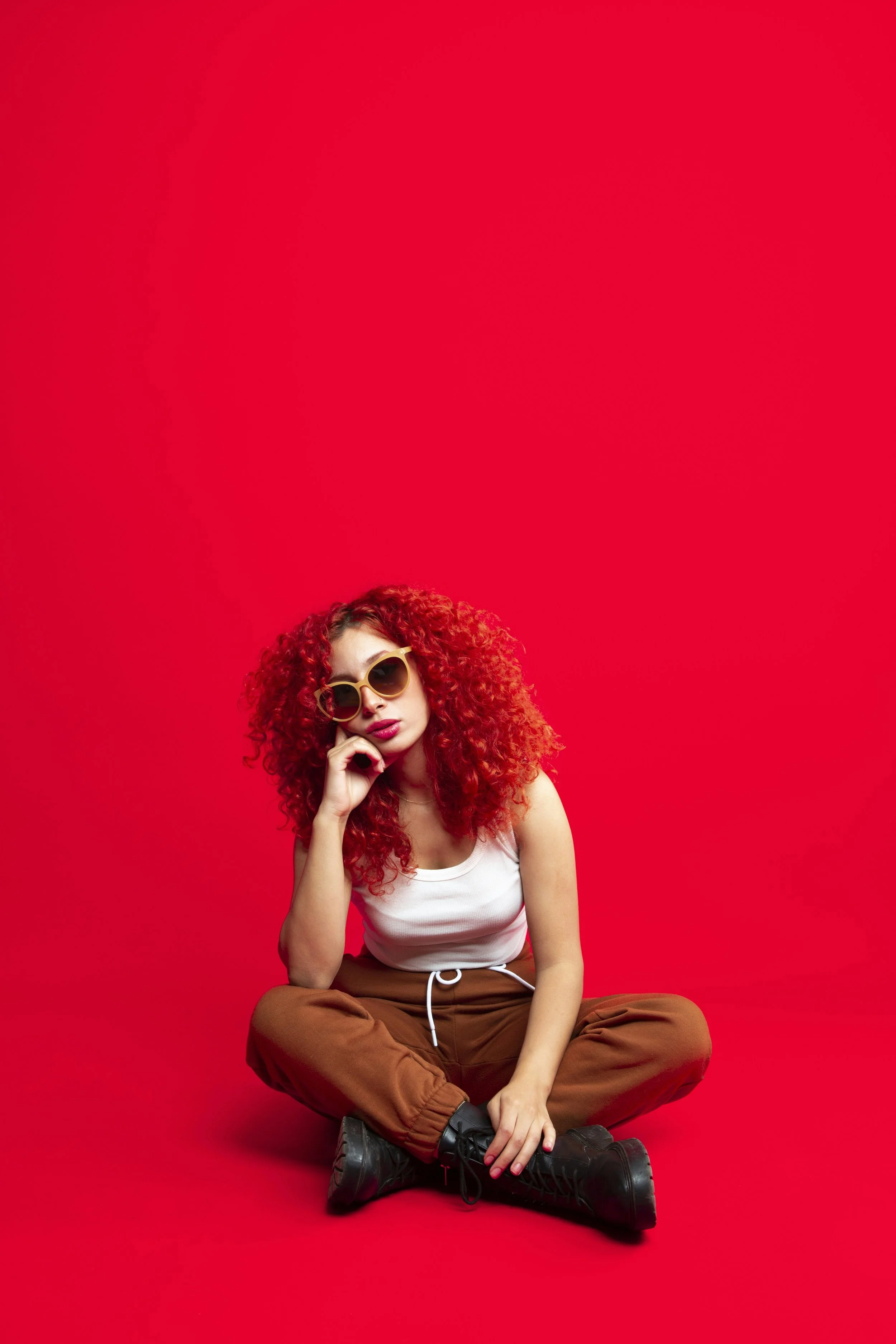 A young woman with curly red hair wearing black sunglasses, a white tank top, brown pants, and black boots, sitting cross-legged against a plain red background.