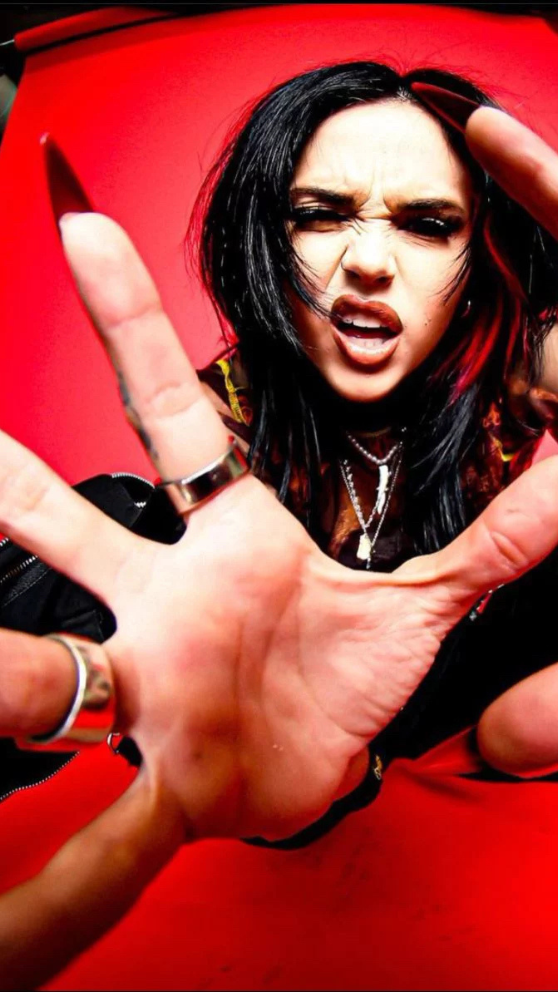 A woman with black hair streaked with red, making a gritty facial expression, reaching into the camera with her hand, with a red background.
