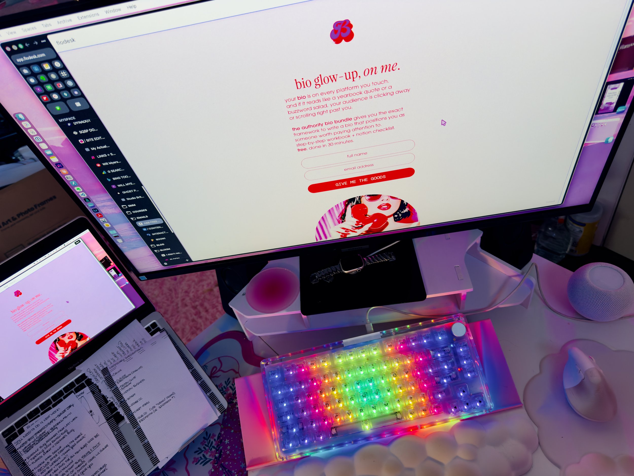 Studio Brittany workspace desk setup showing a Flodesk landing page for the Authority Bio Bundle on a monitor, with a rainbow LED mechanical keyboard, iPad displaying a pink website, handwritten notes, and pink and purple ambient lighting.
