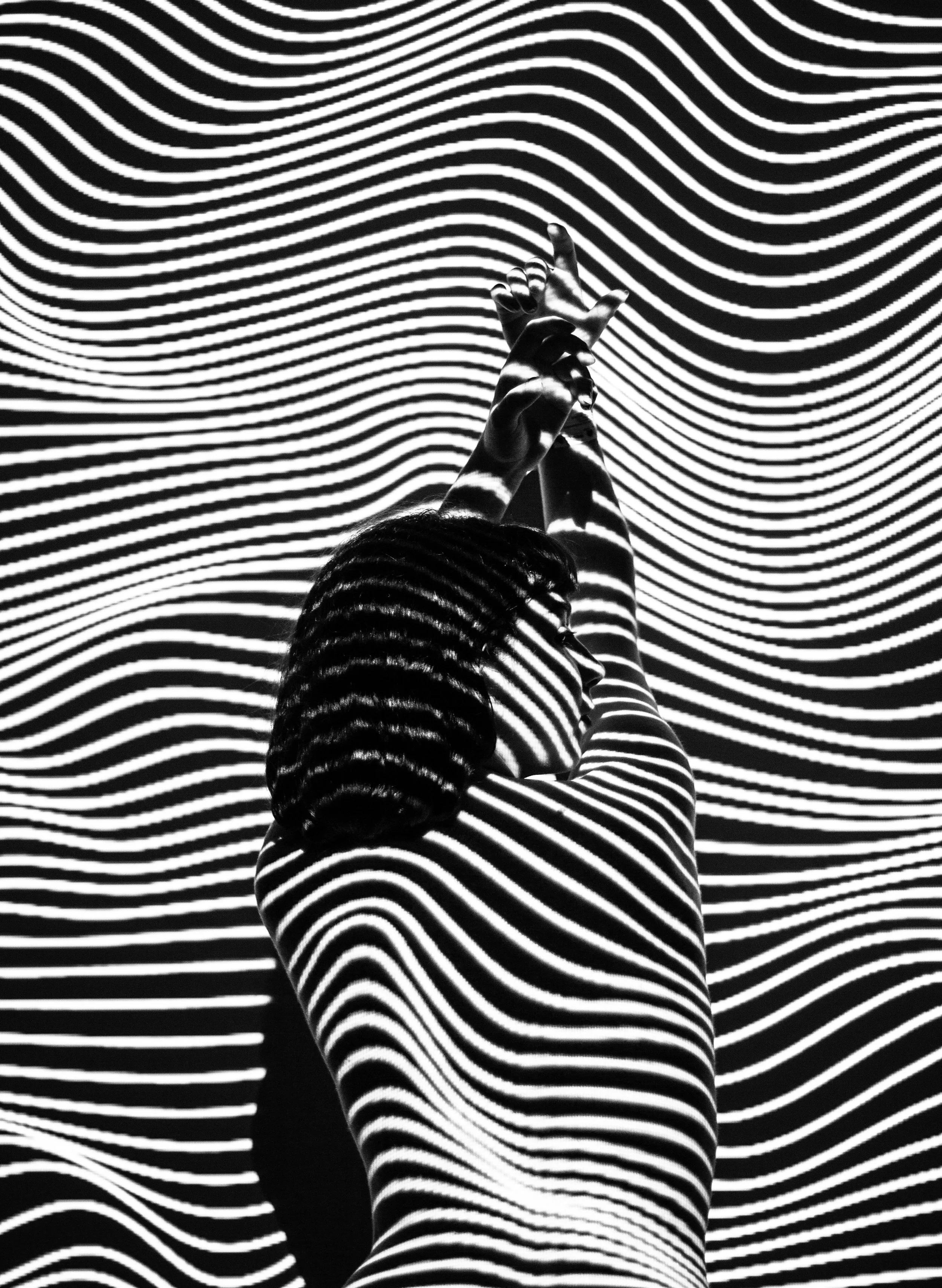 A person with long hair, wearing striped clothing, is reaching up with both hands, with black and white hypnotic circle patterns projected onto their body and the background.