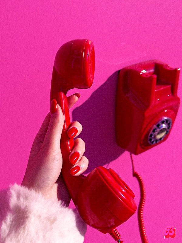 A red phone held by a manicured hand with red nails with a bright hot pink/magenta background.