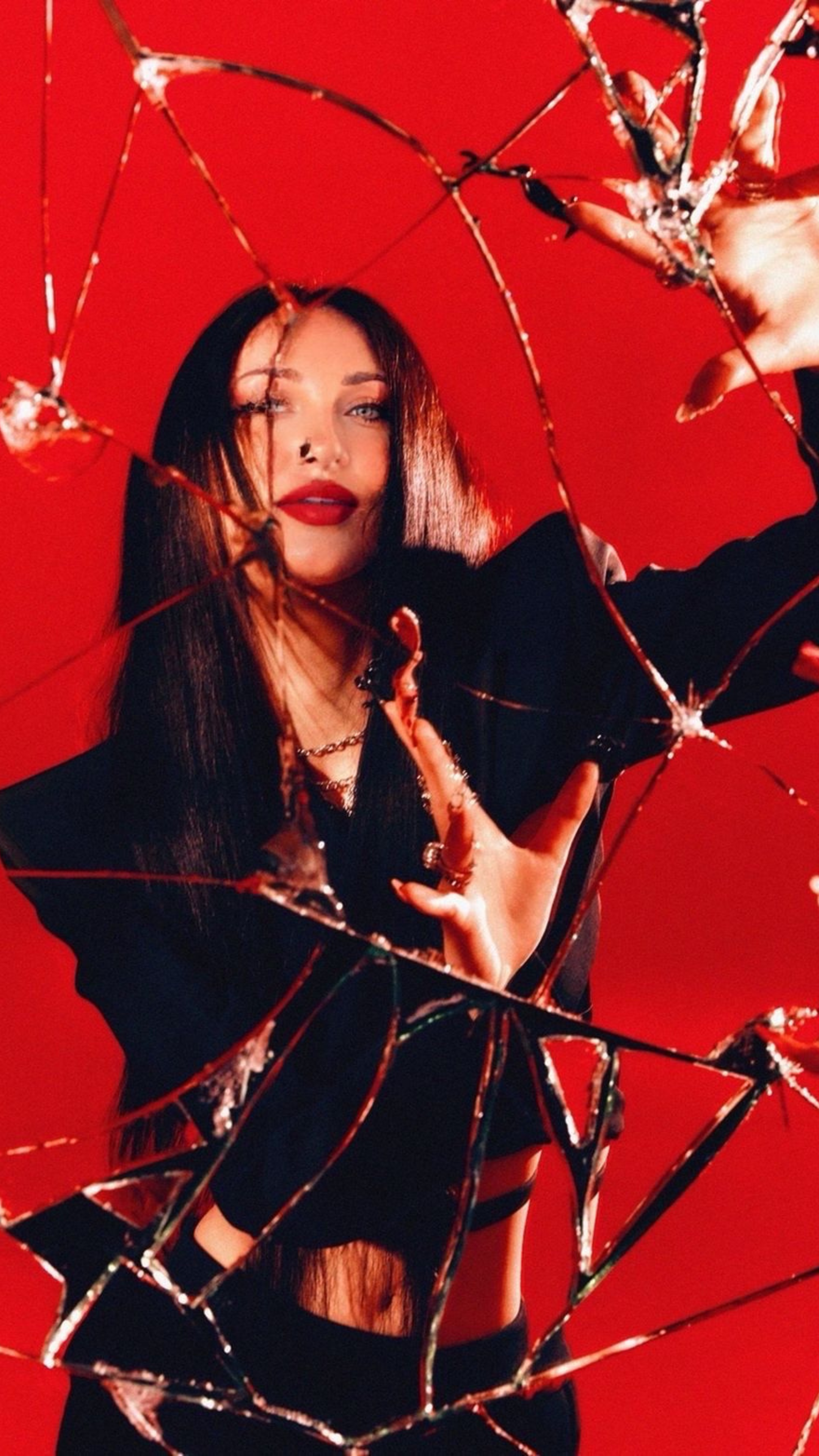 A woman with long dark hair, wearing dark clothing and red lipstick, is seen behind a broken mirror with fragmented reflections, against a solid red background.