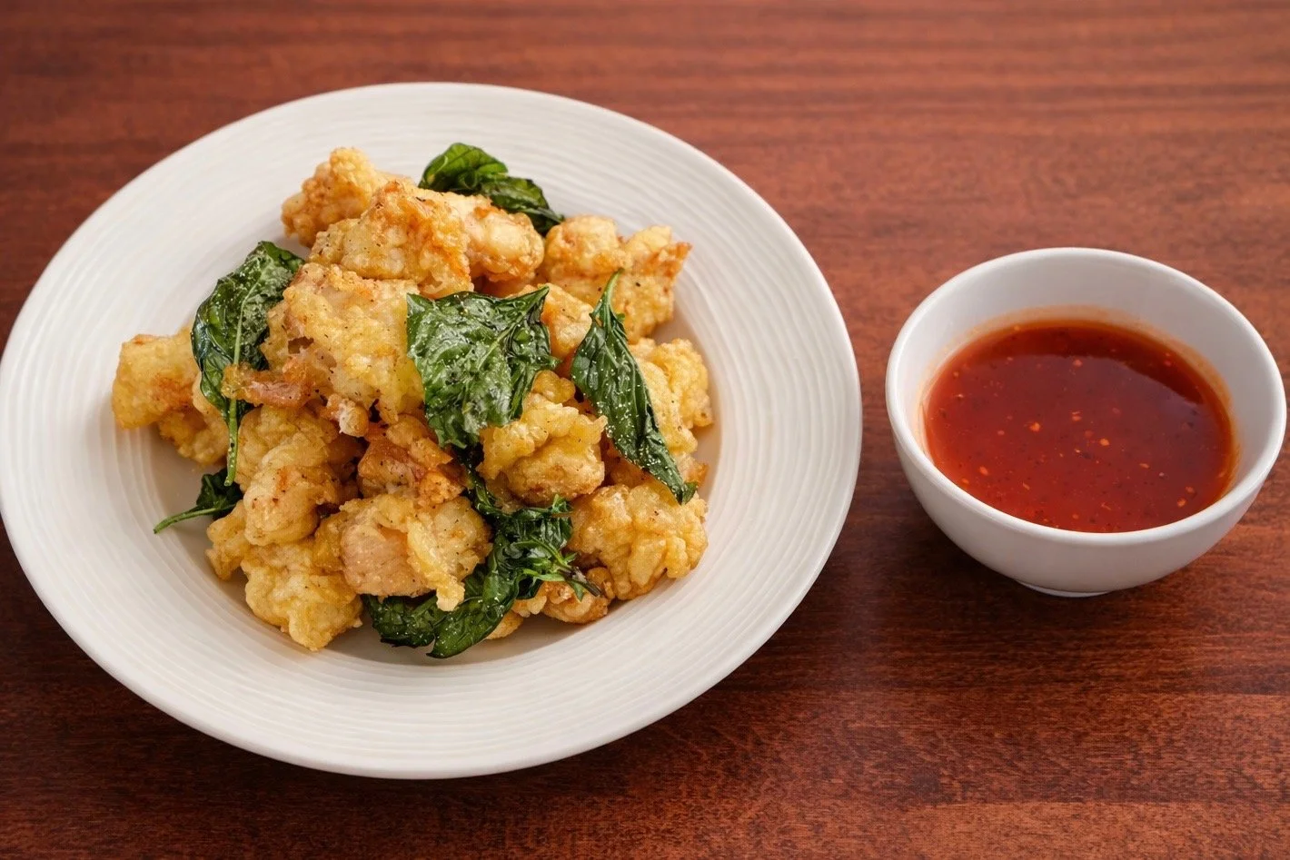 Fried chicken pieces with green leafy herbs on a white plate, served with a small white bowl of red dipping sauce on a wooden table.