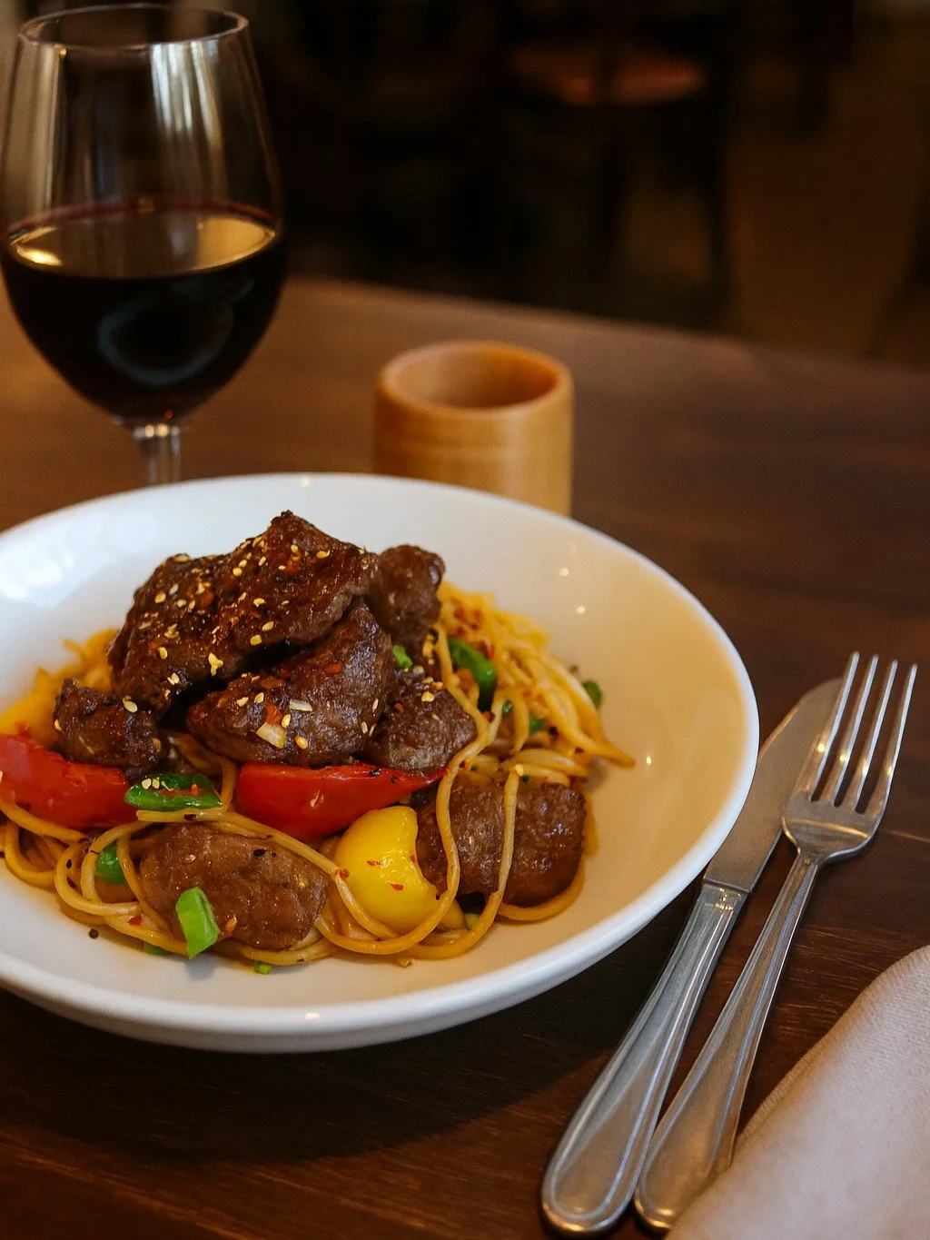 Black Pepper Steak with Noodles (Bò Xào Tiêu Đen) Seared juicy and tender beef fillet with noodles and peppers in a bold, garlic butter, and fragrant black pepper sauce. A fusion of Vietnamese, served with a glass of red wine on a wooden table.