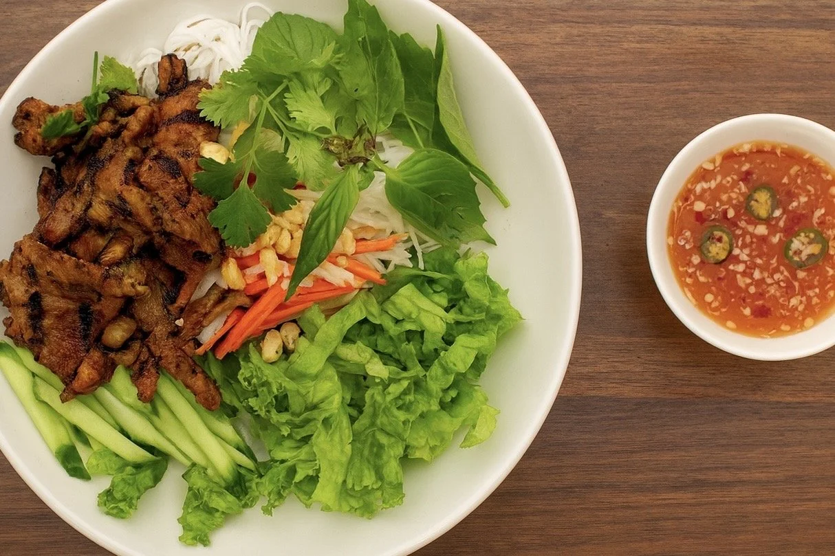 A bowl of grilled meat, fresh greens, shredded vegetables, and rice noodles with a side of spicy dipping sauce.