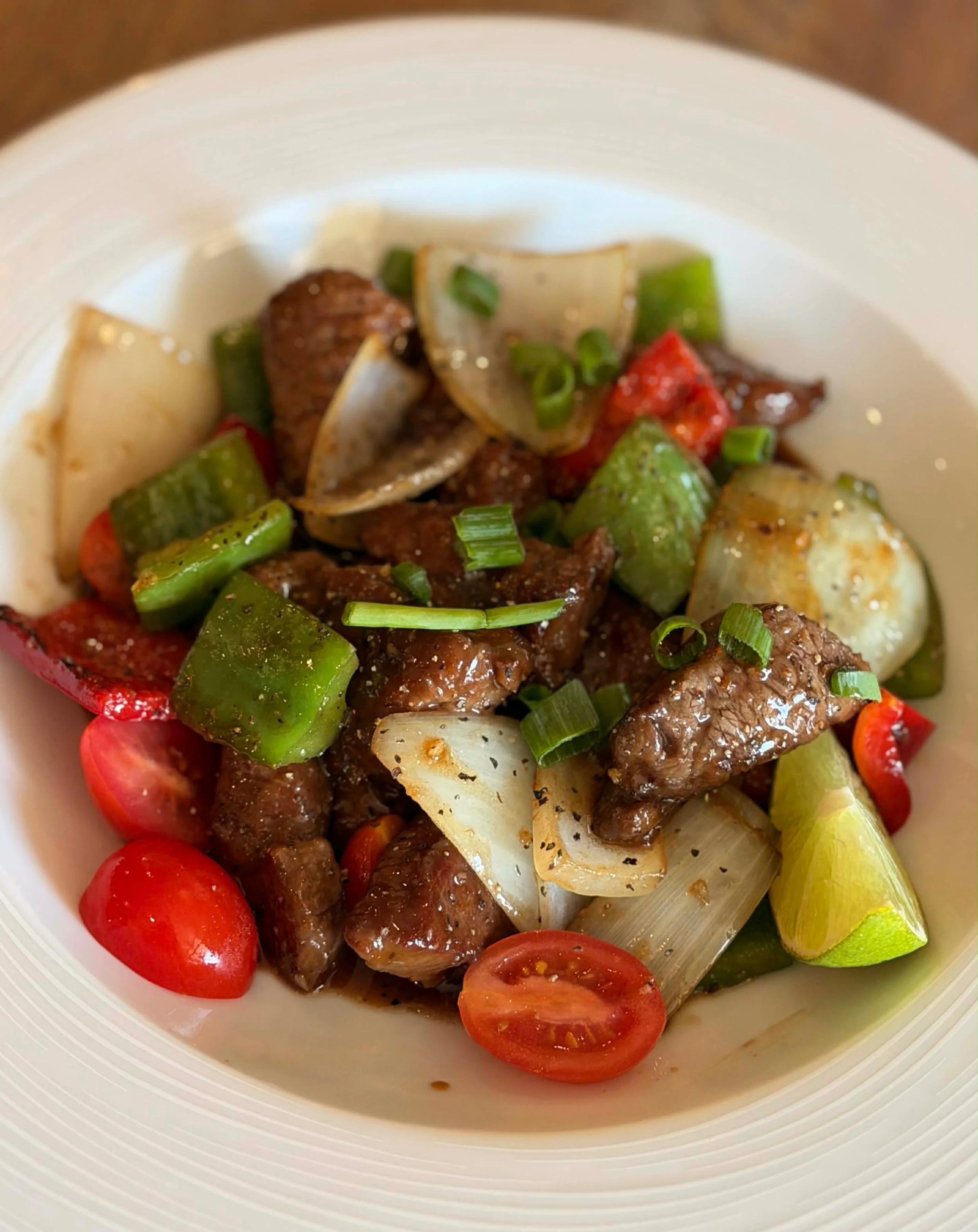 A bowl of stir-fried beef with cherry tomatoes, green and white onions, and green bell peppers, garnished with chopped green onions.