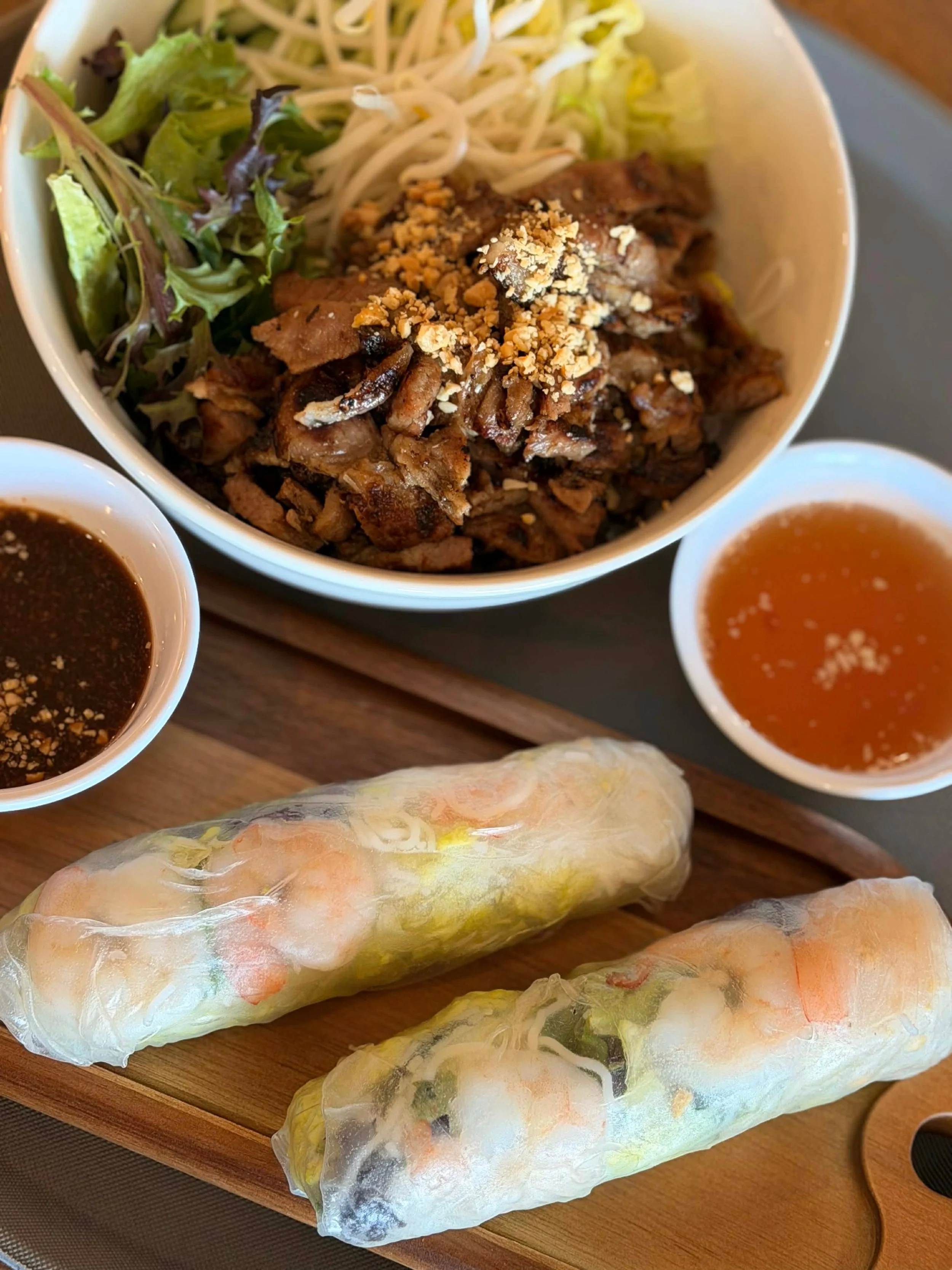 Vietnamese cuisine featuring two fresh spring rolls with shrimp and vegetables, a bowl of salad with grilled meat topped with crushed peanuts, and two dipping sauces, one dark and one orange, all served on a wooden tray.