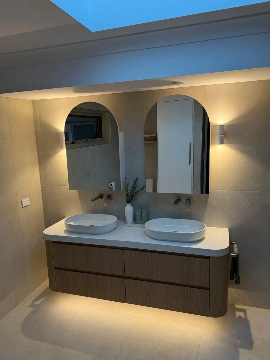 Modern bathroom with two vessel sinks on a wooden vanity, large mirror, beige tiled walls, wall sconces, and a skylight ceiling.