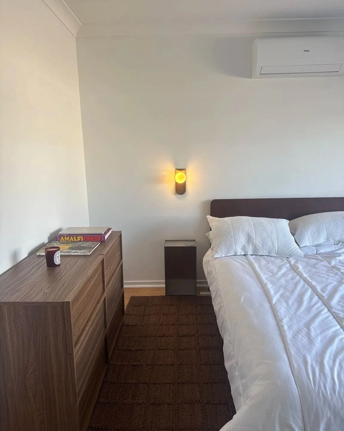 Hotel bedroom with a white bed, pillows, a bedside table, wall-mounted light, a wooden dresser with magazines and a candle, and an air conditioning unit on the wall.
