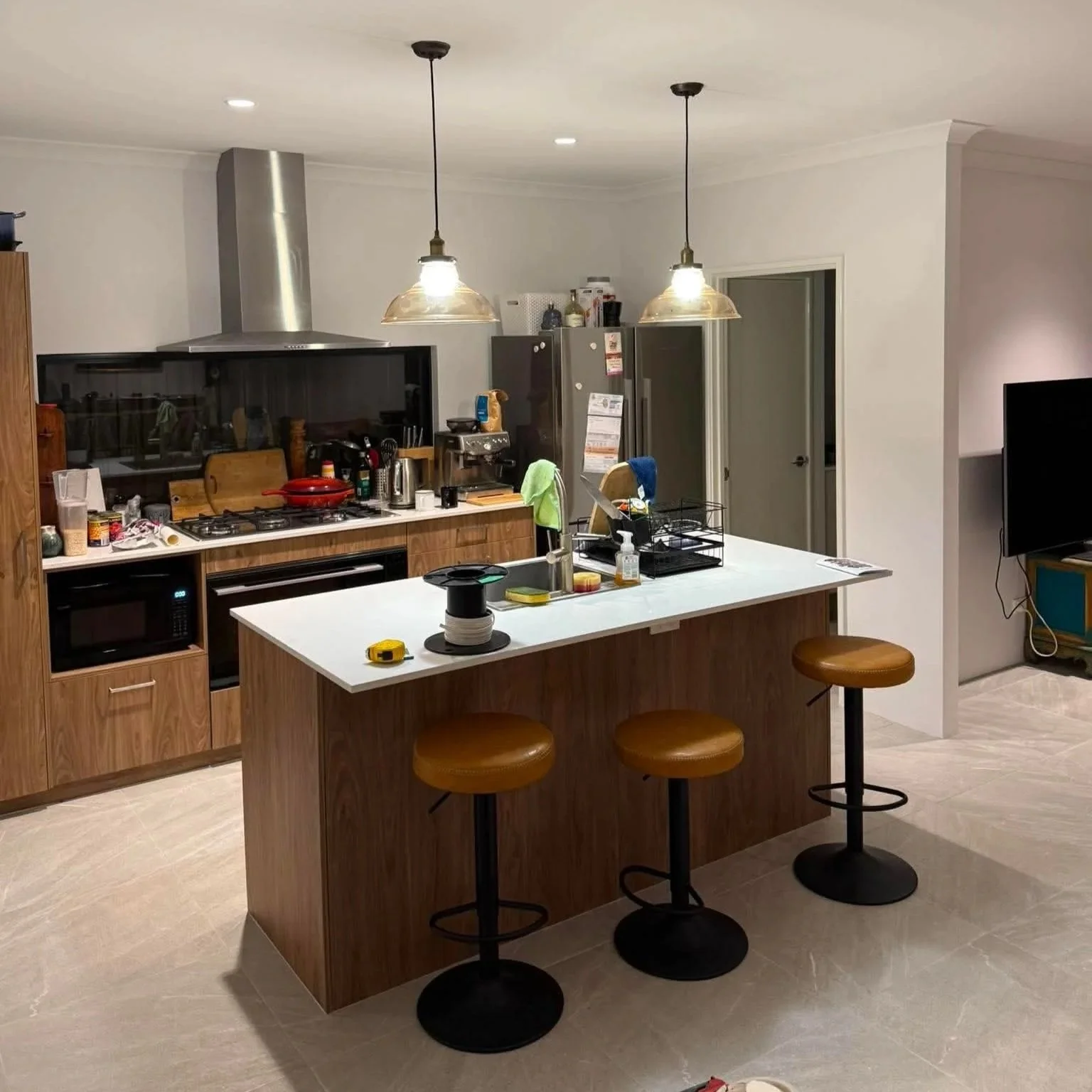 Kitchen with white island counter, four brown bar stools, wooden cabinets, stainless steel stove with range hood, small microwave, refrigerator, and various kitchen items, in a modern home.