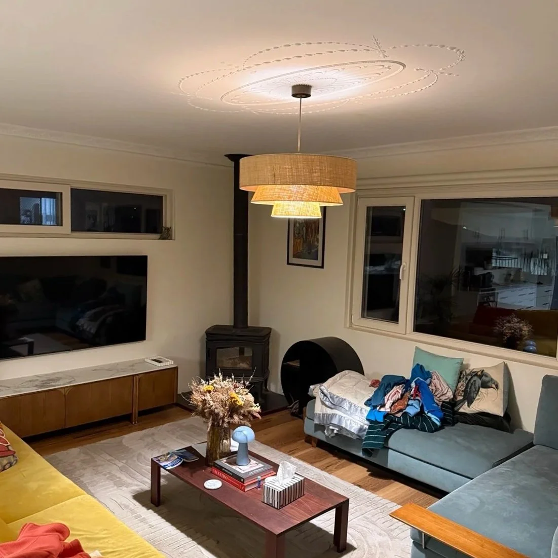Living room with a TV, a wood-burning stove, a hanging light fixture, couches, a coffee table with books, a vase with flowers, and clothes on the couch.