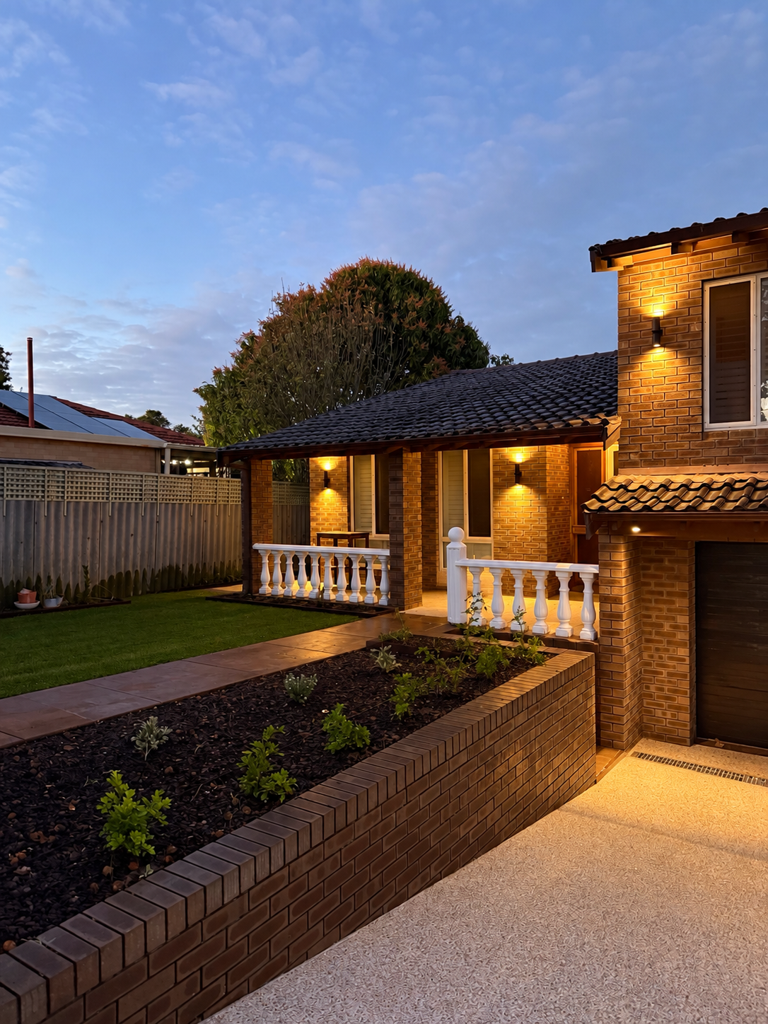 A brick house exterior at dusk with illuminated outdoor lights on the porch and walls, a tiled walkway, a garden bed with plants, a grassy yard, and a fence with solar panels on neighboring roof in the background.