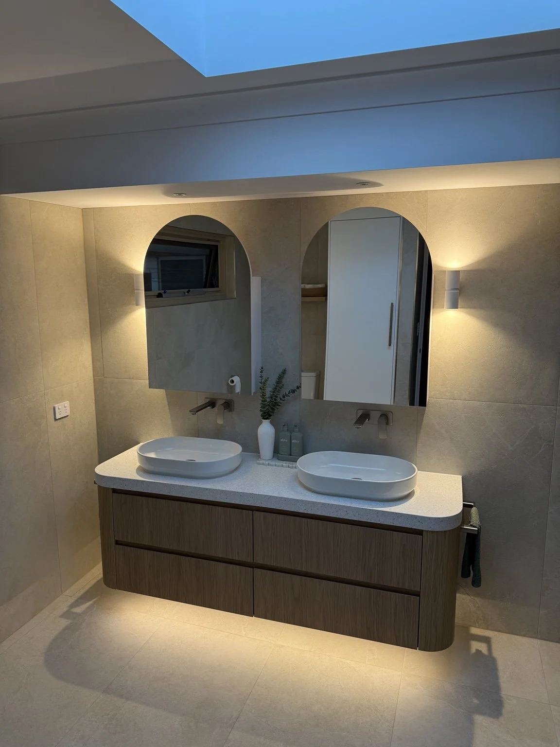 Modern bathroom with double oval vessel sinks on a wooden vanity, large mirror with two wall sconces, small window, and light-colored tiled walls and floor.