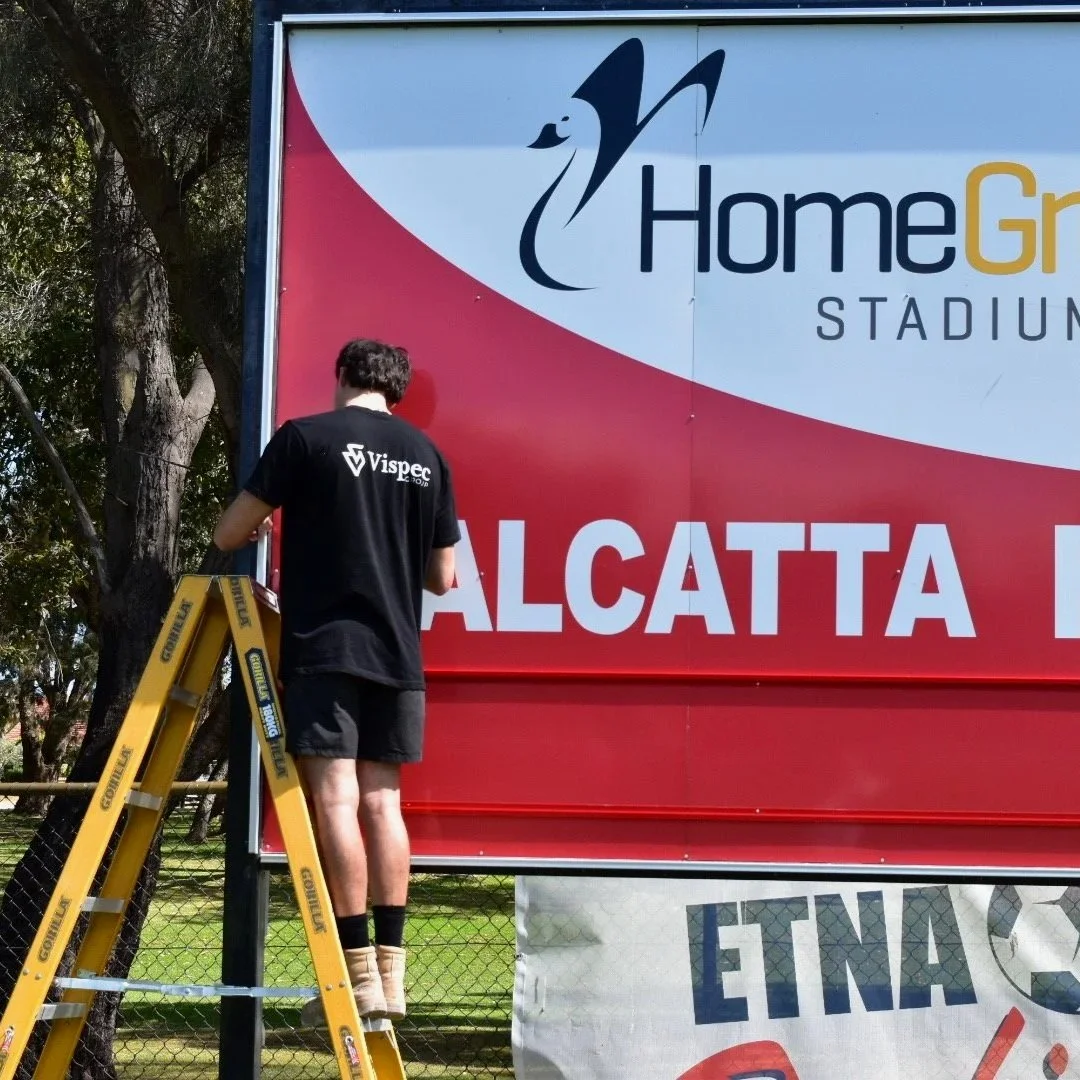 A person standing on a yellow ladder installing a led lighting system or repairing a sign for Home Group Stadium. The sign has a partial logo with a stylized wolf or fox and text, with trees in the background.