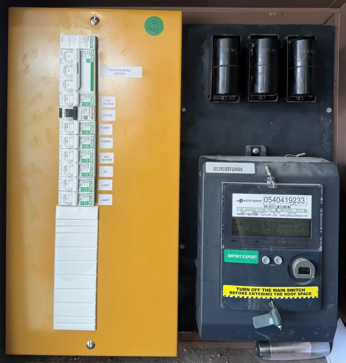 Electrical control panel with labeled switches and a digital meter, installed on a wall, with a caution sign indicating to turn off the main switch before entering the roof space.