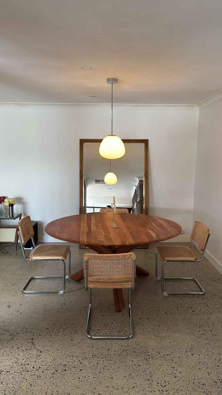 Indoor dining area with a round wooden table, four wicker chairs, a large rectangular mirror on the wall, hanging pendant lights, and a small side table with items on it.