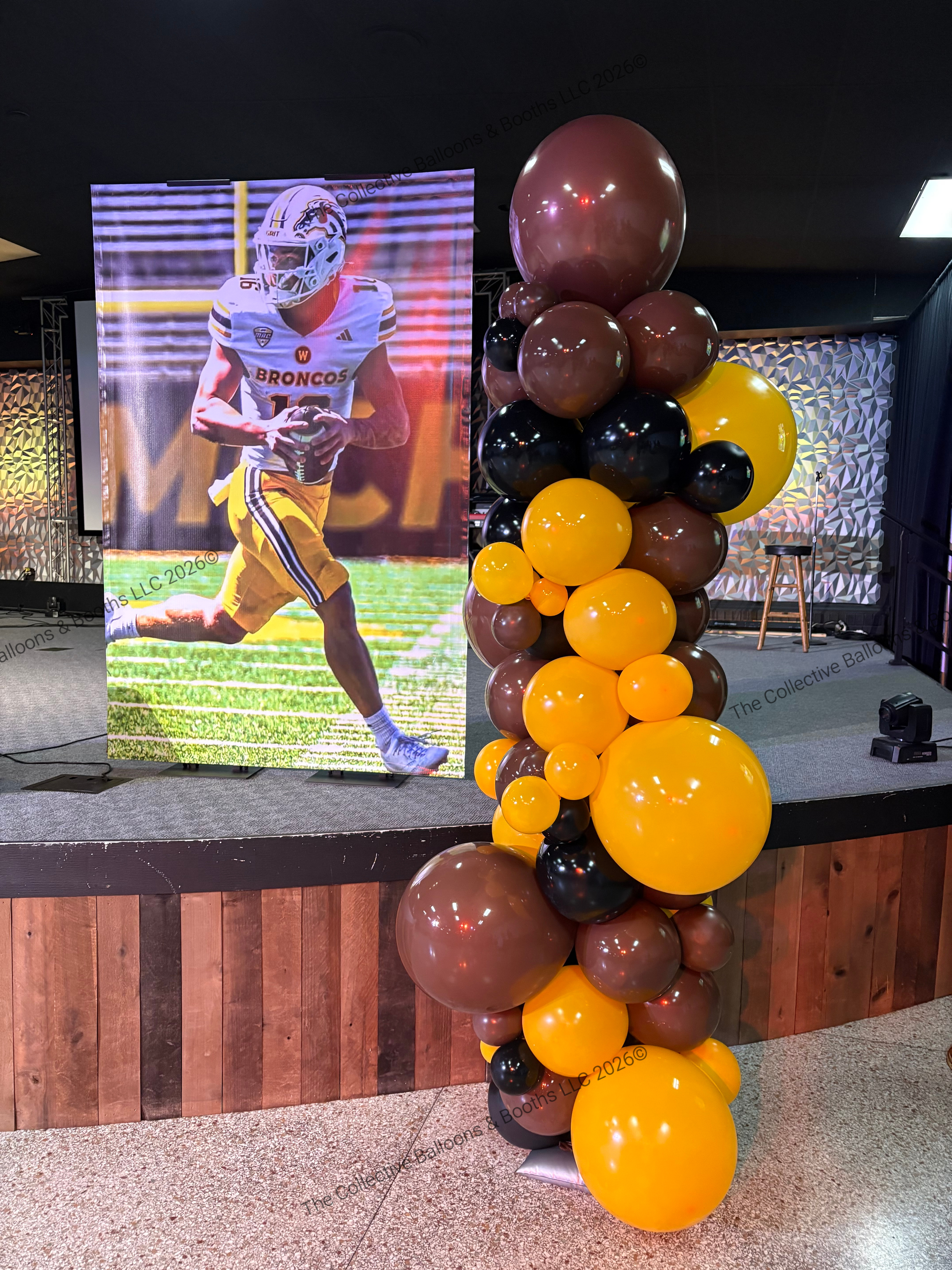 A large display featuring a football player in a Broncos uniform, standing on a stage decorated with a cluster of black, yellow, and brown balloons, and a stool in the background.
