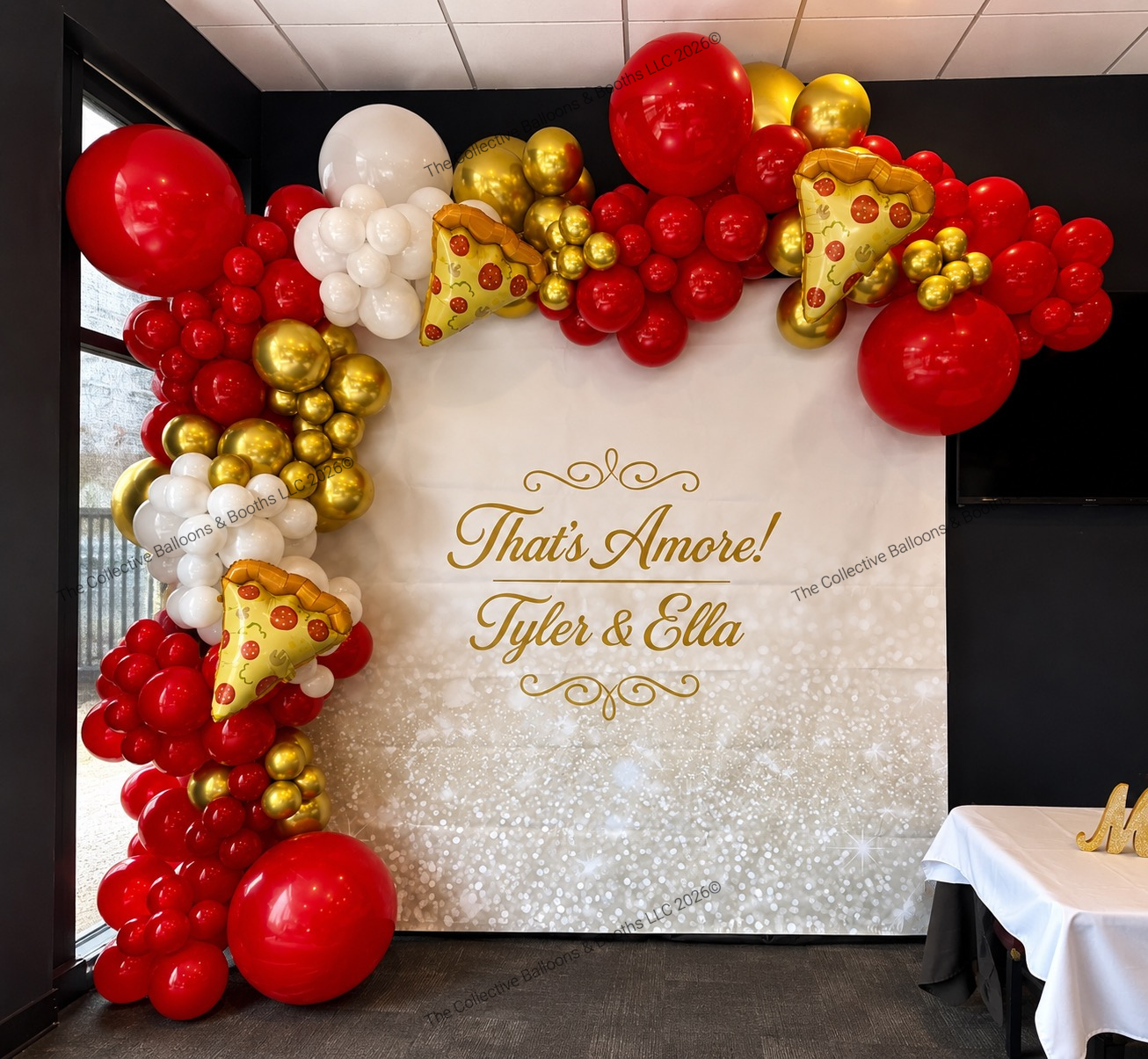 Balloon arch with red, white, and gold balloons framing a white backdrop with gold lettering that says "That's Amore! Tyler & Ella" at a celebration or wedding event.