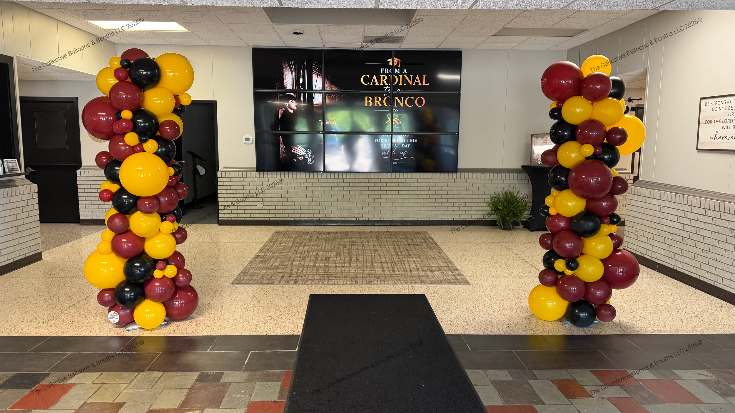 Balloon decorations with maroon, black, and yellow balloons on either side of an entrance in a building lobby, leading to a digital display board with the words 'From a Cardinal to a Bronc.'