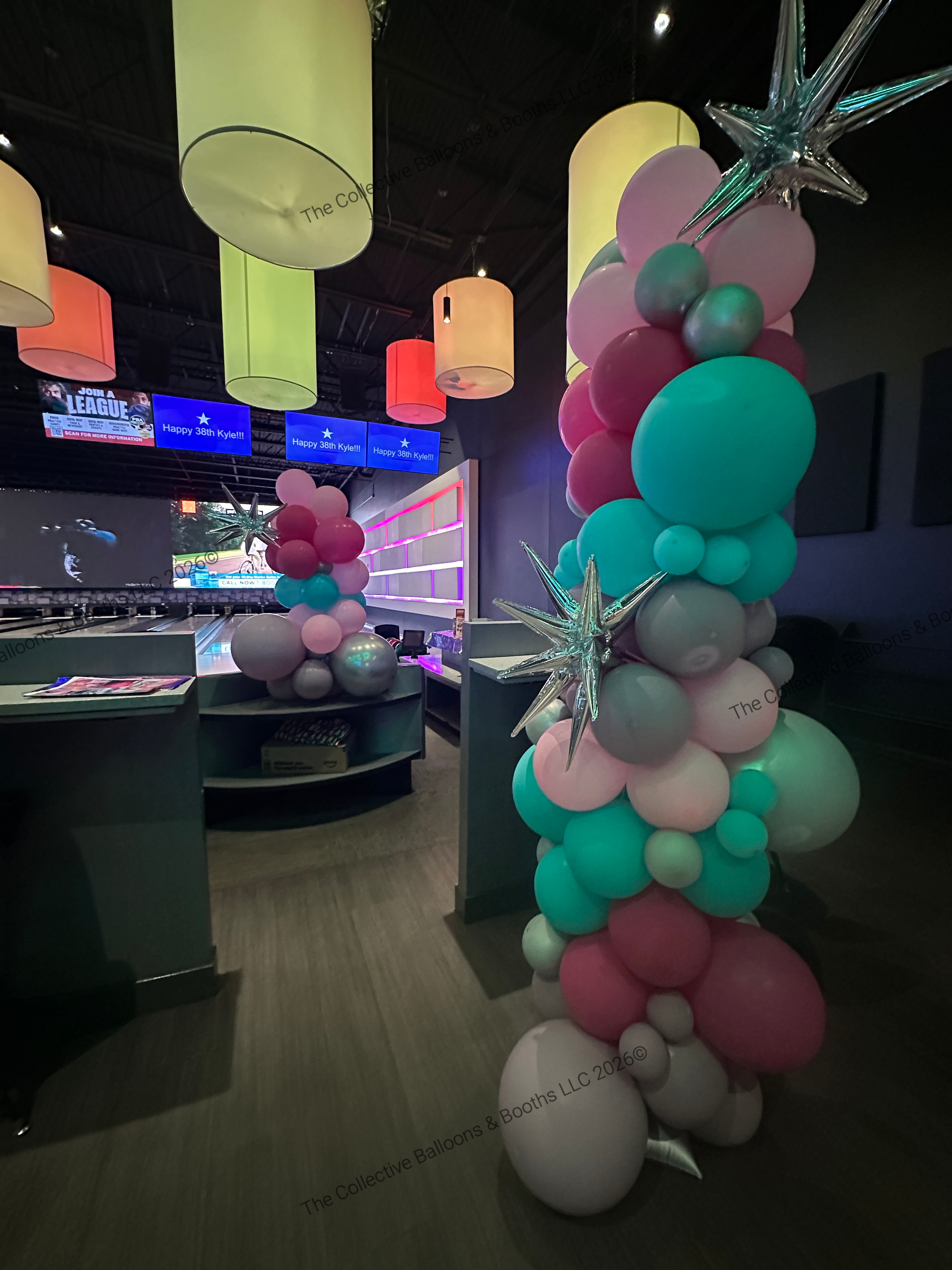 Vertical balloon column with pink, teal, white, and silver balloons, topped with silver star-shaped balloons, inside a room with hanging colored lamps and digital screens.