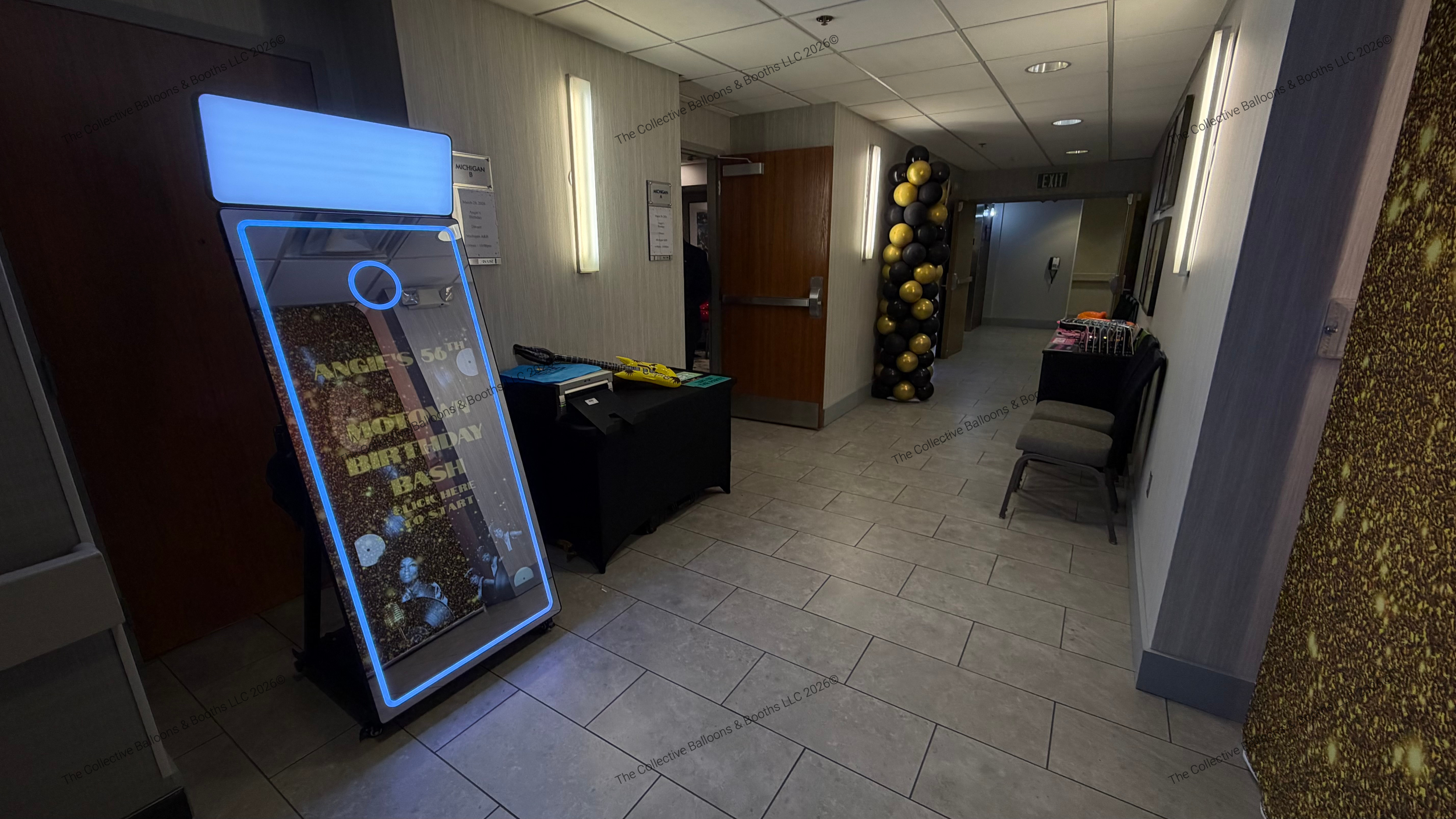 A hallway with chairs against the wall, a tall column of black and yellow balloons, a neon sign promoting a birthday bash, and a small table with items including a toy helicopter. The hallway has beige walls, tile flooring, and fluorescent lighting.