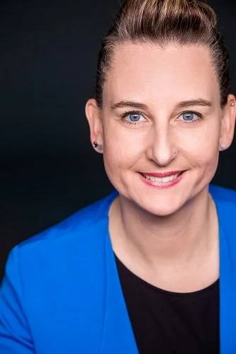 Lächelnde Frau mit braunen Haaren in einem Dutt, trägt orangefarbenen Blazer und blauen Oberteil, im Innenraum.