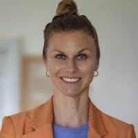 Lächelnde Frau mit braunen Haaren in einem Dutt, trägt orangefarbenen Blazer und blauen Oberteil, im Innenraum.