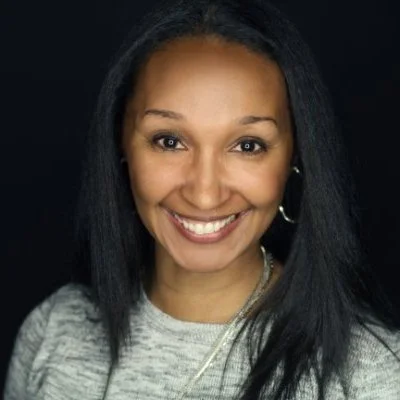 A smiling woman with long black hair, hoop earrings, and wearing a light gray top against a dark background.