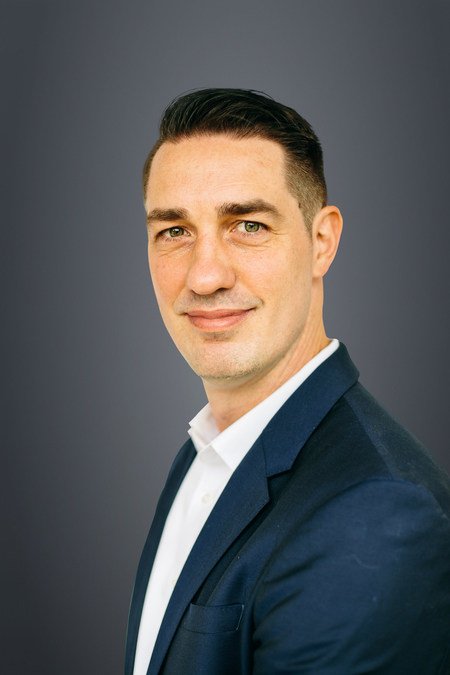 A professional man wearing a navy blazer and white shirt, with dark hair neatly combed, smiling subtly against a gray background.