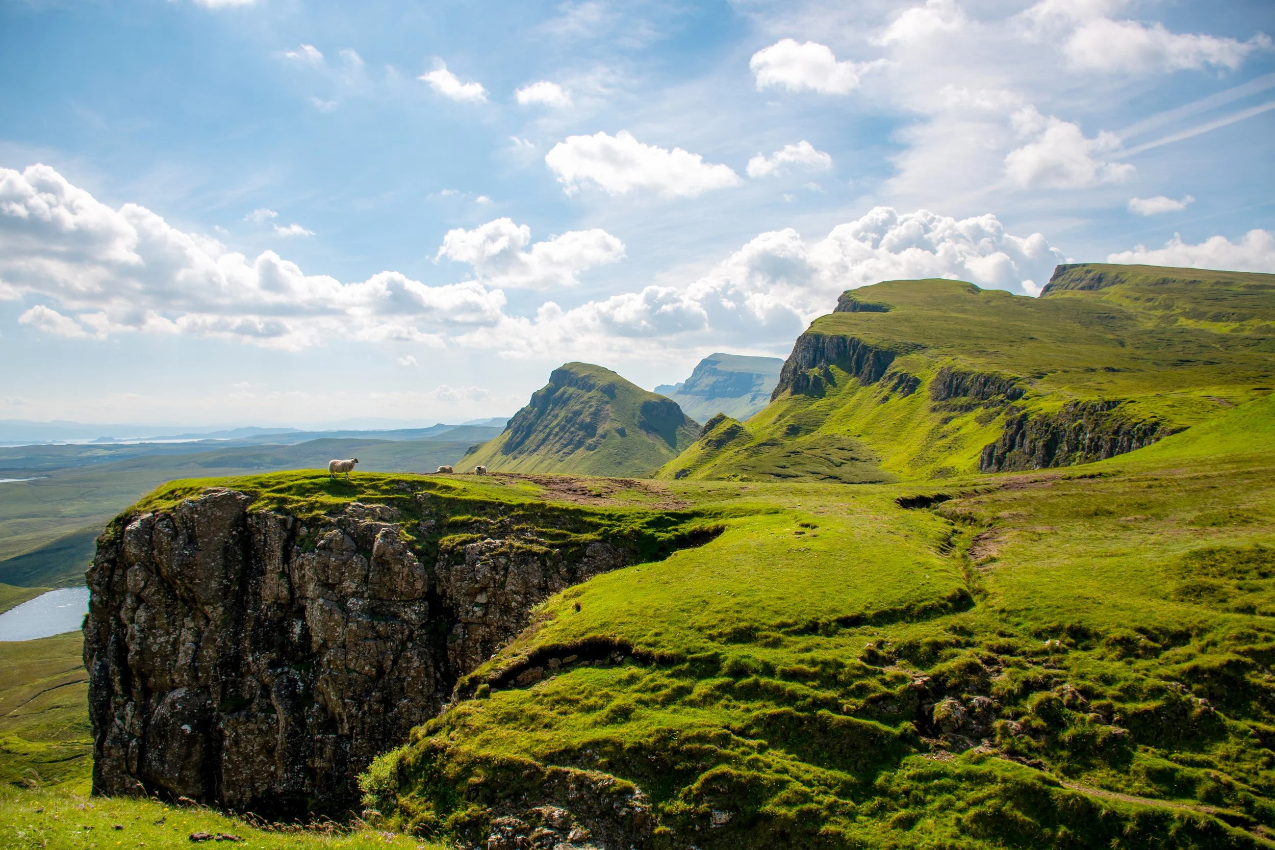 Green rolling hills and cliffs under a partly cloudy sky, with sheep grazing on the grassy area.