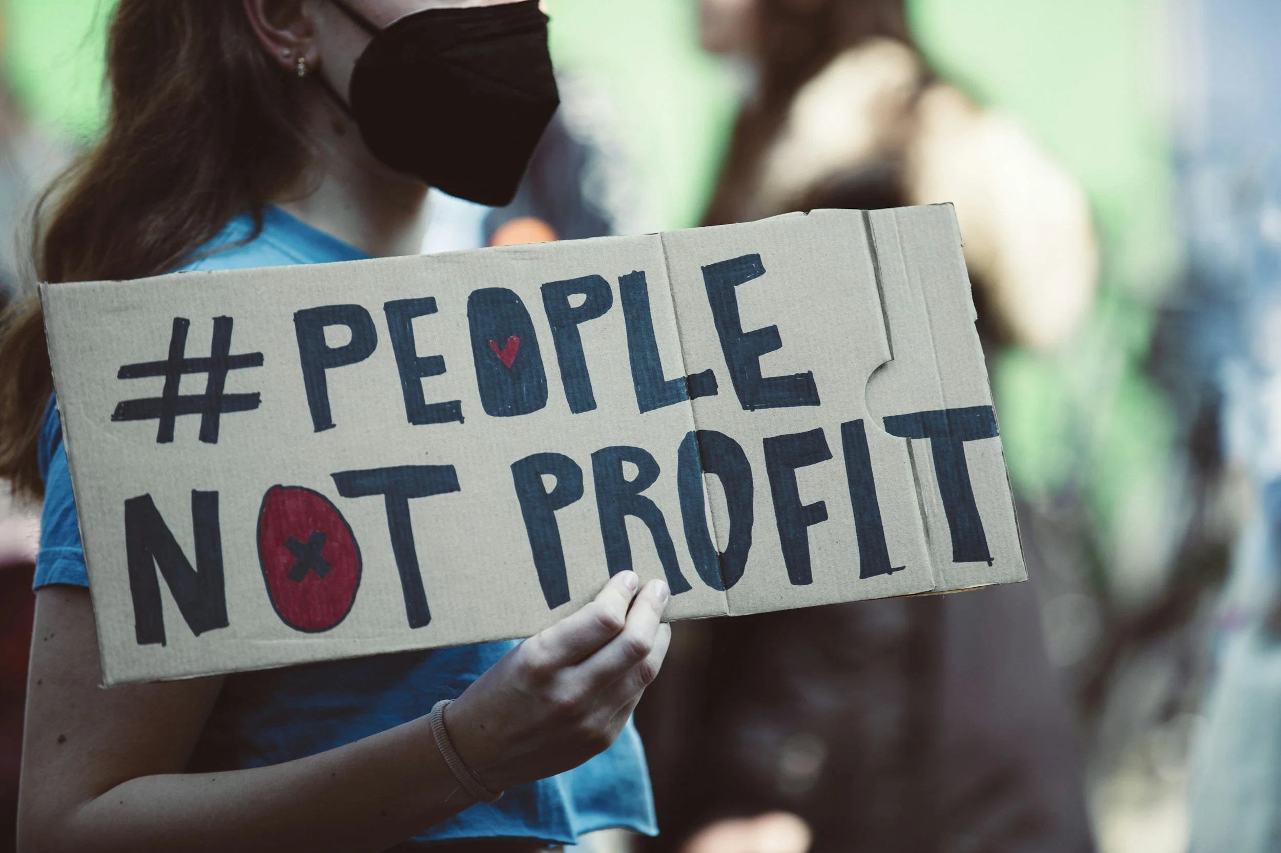 Person wearing a black face mask holding a cardboard sign that reads '#PEOPLE NOT PROFIT' with a red heart and circle symbols, in a crowd.