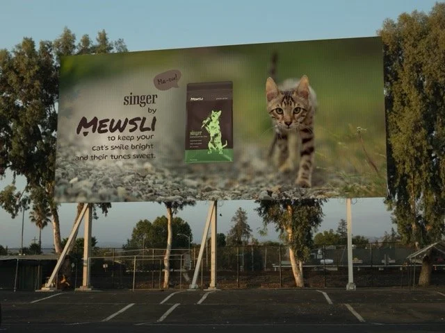 Large billboard advertising 'Singer by Mewsli' cat food featuring a kitten on a grassy outdoor background.