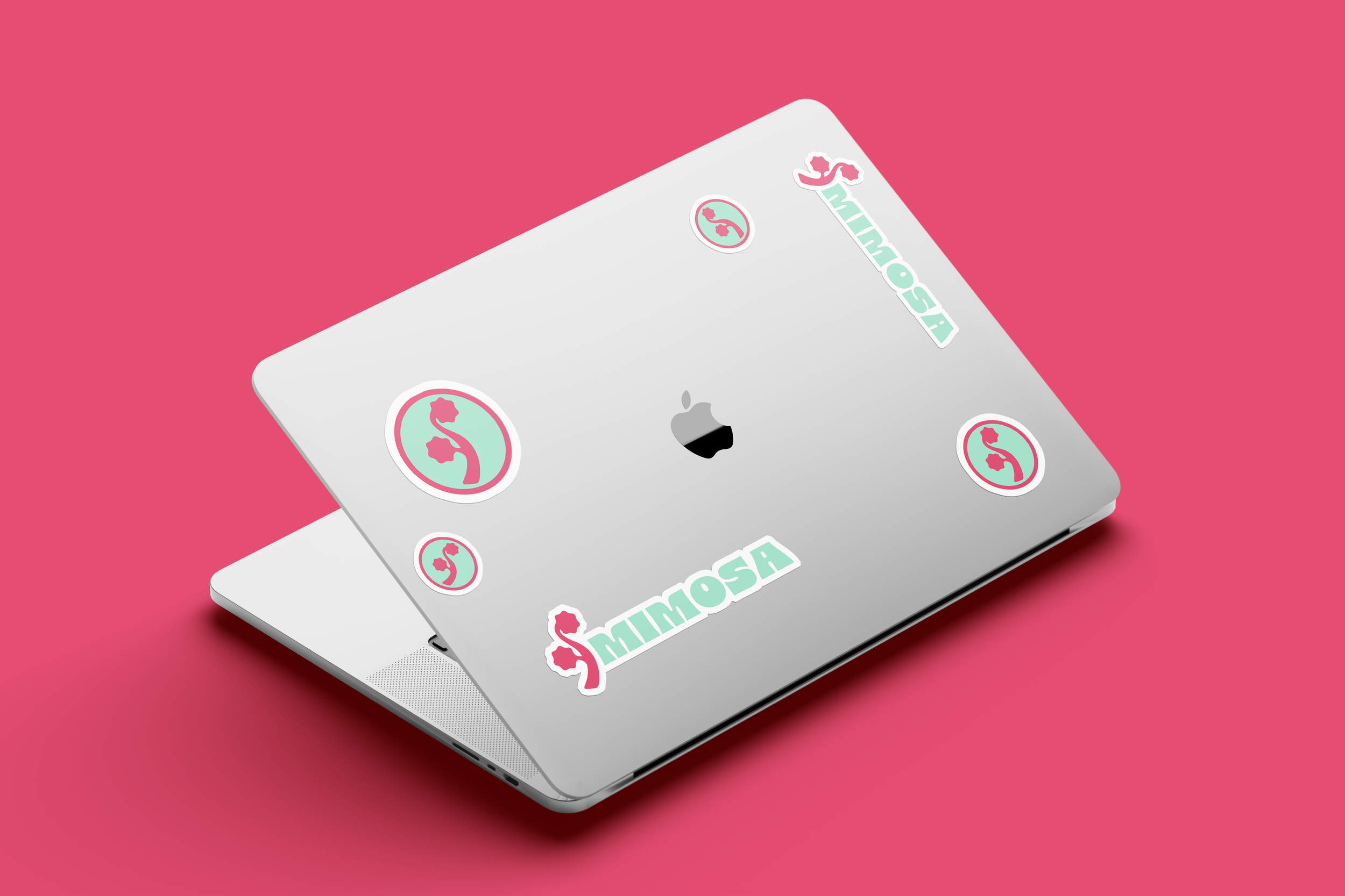 A silver MacBook laptop with pink and green stickers on the lid, placed on a pink background. The stickers include a stylized animal logo and the word 'MIMOSA'.