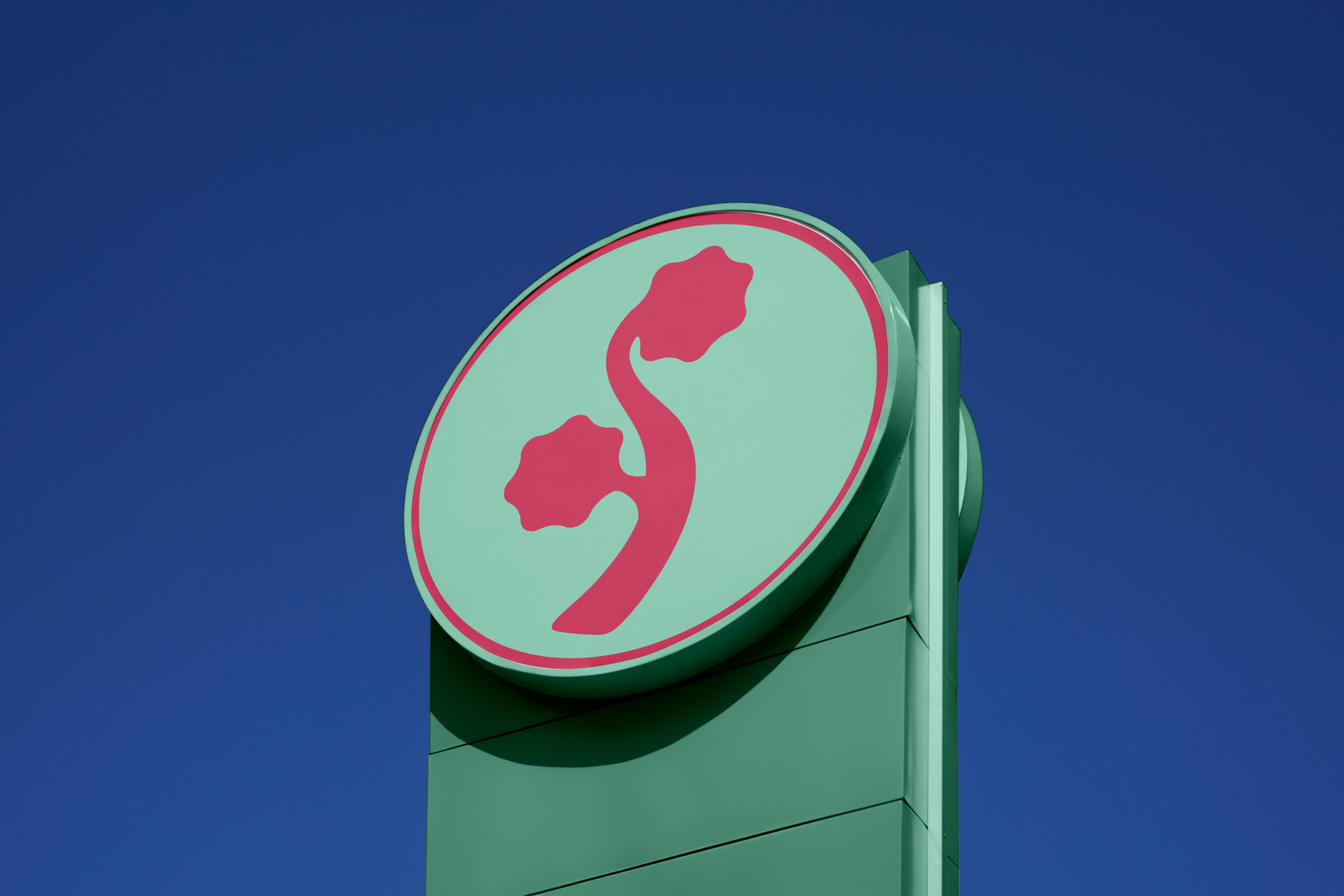 A round sign with a stylized pink flower logo on a light green background, mounted on a green pole against a clear blue sky.