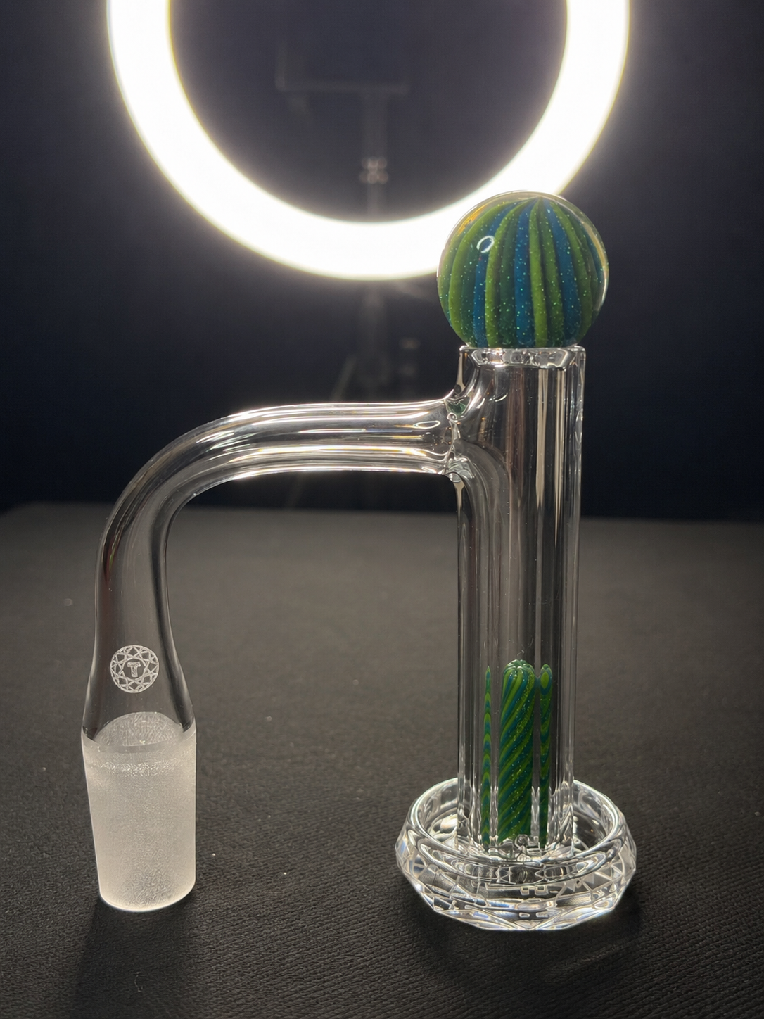 Glass water pipe with a marble on top and decorative green and blue elements inside, placed on a black surface with a bright ring light in the background.