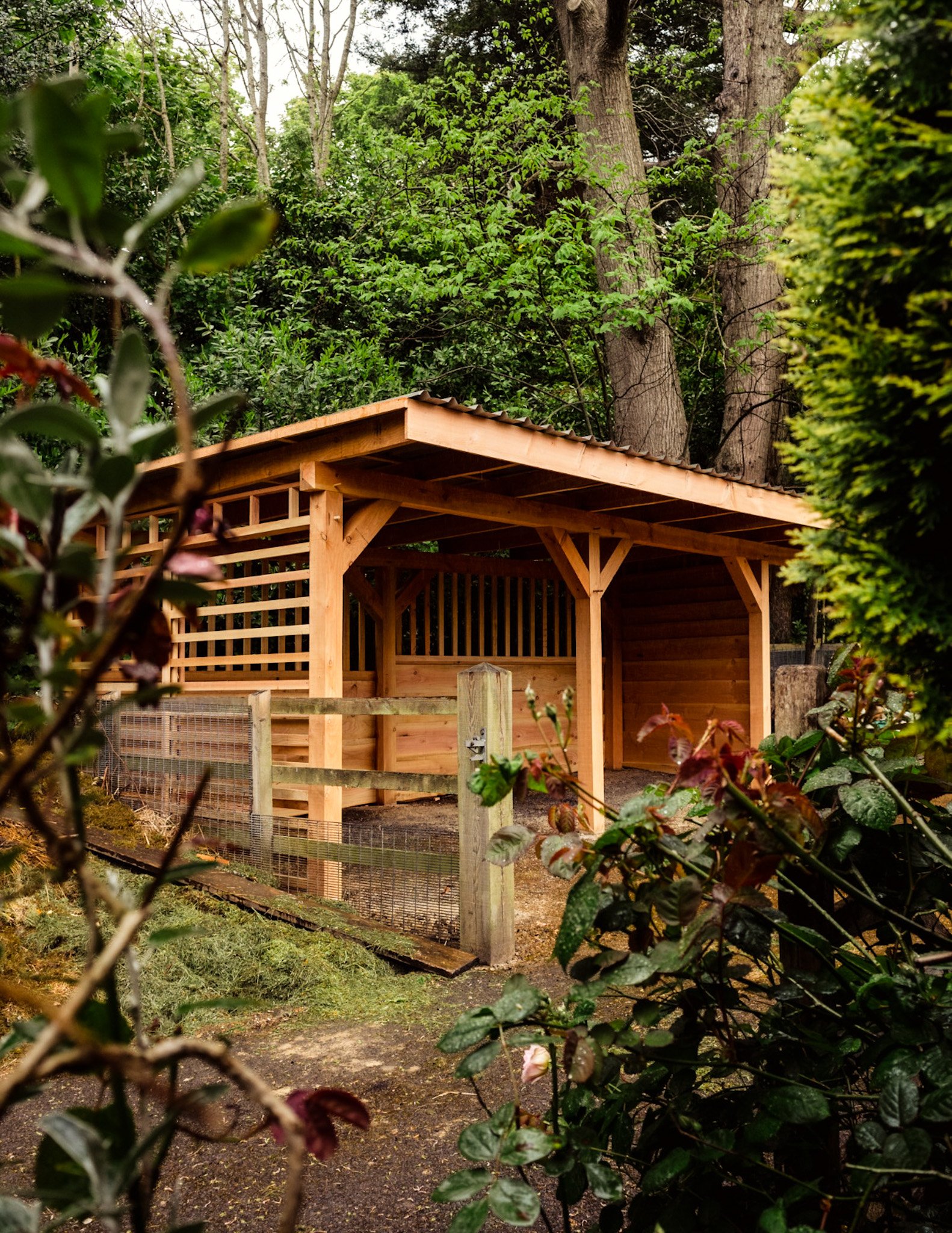 A wooden garden shed with a sloped roof, situated among green trees and shrubs, with a wooden fence and a small gate in the foreground.