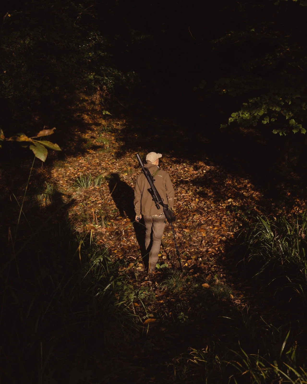 A person wearing outdoor gear walking through a dark, wooded area at night, carrying a rifle on their shoulder.