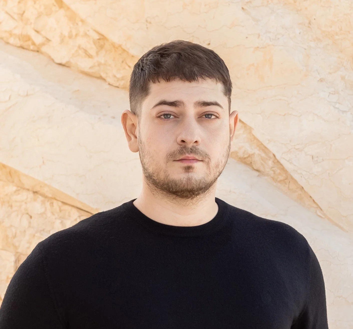 A young man with short dark hair, light skin, and facial hair wearing a black sweater, standing in front of a beige stone wall with textured patterns.