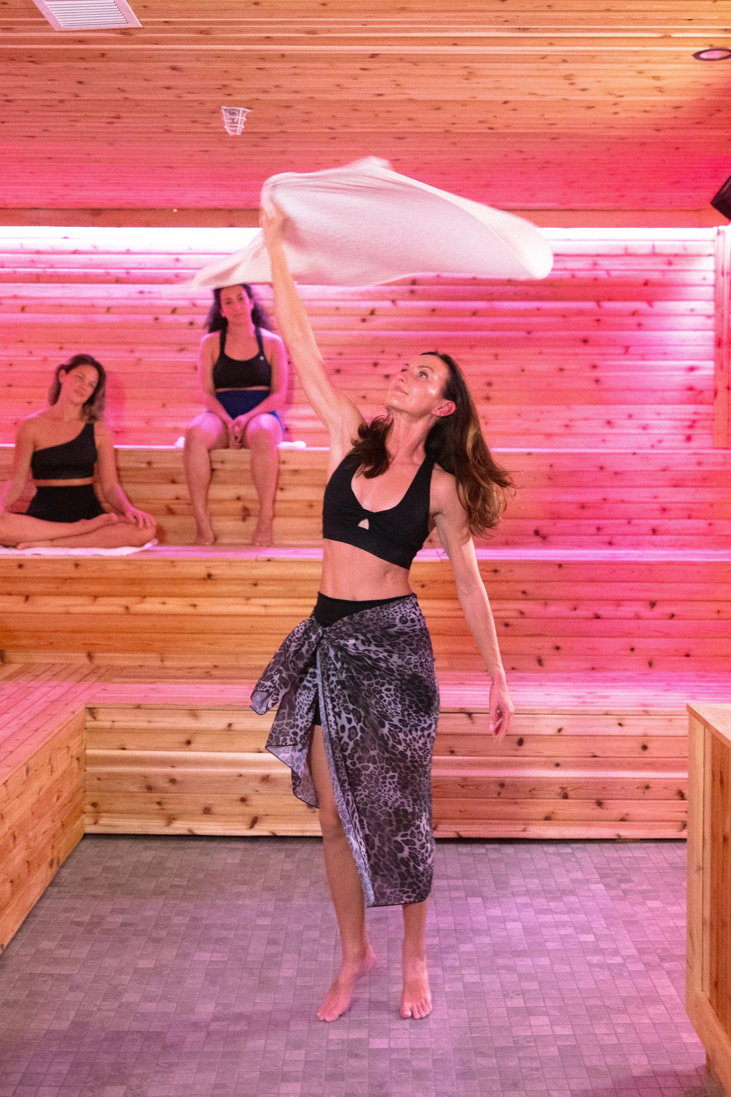 A woman in a black sports bra and leopard-print skirt is performing a dance move with a white cloth in a yoga or dance studio with pink lighting. Two women sitting on wooden benches are watching her.