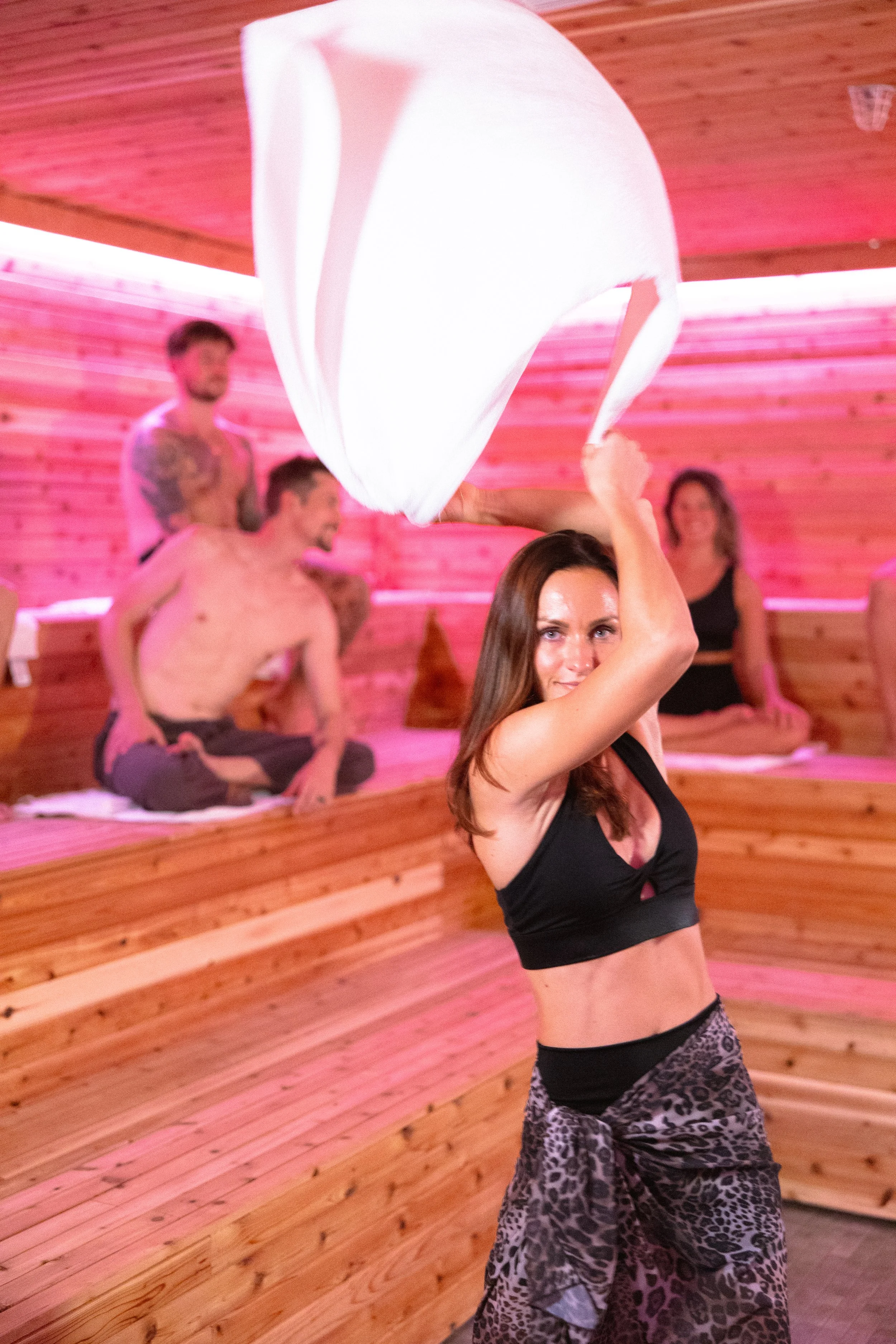 Woman doing a yoga pose with a white cloth in a wooden room with pink pink lighting, while other men and women sit and relax in the background.
