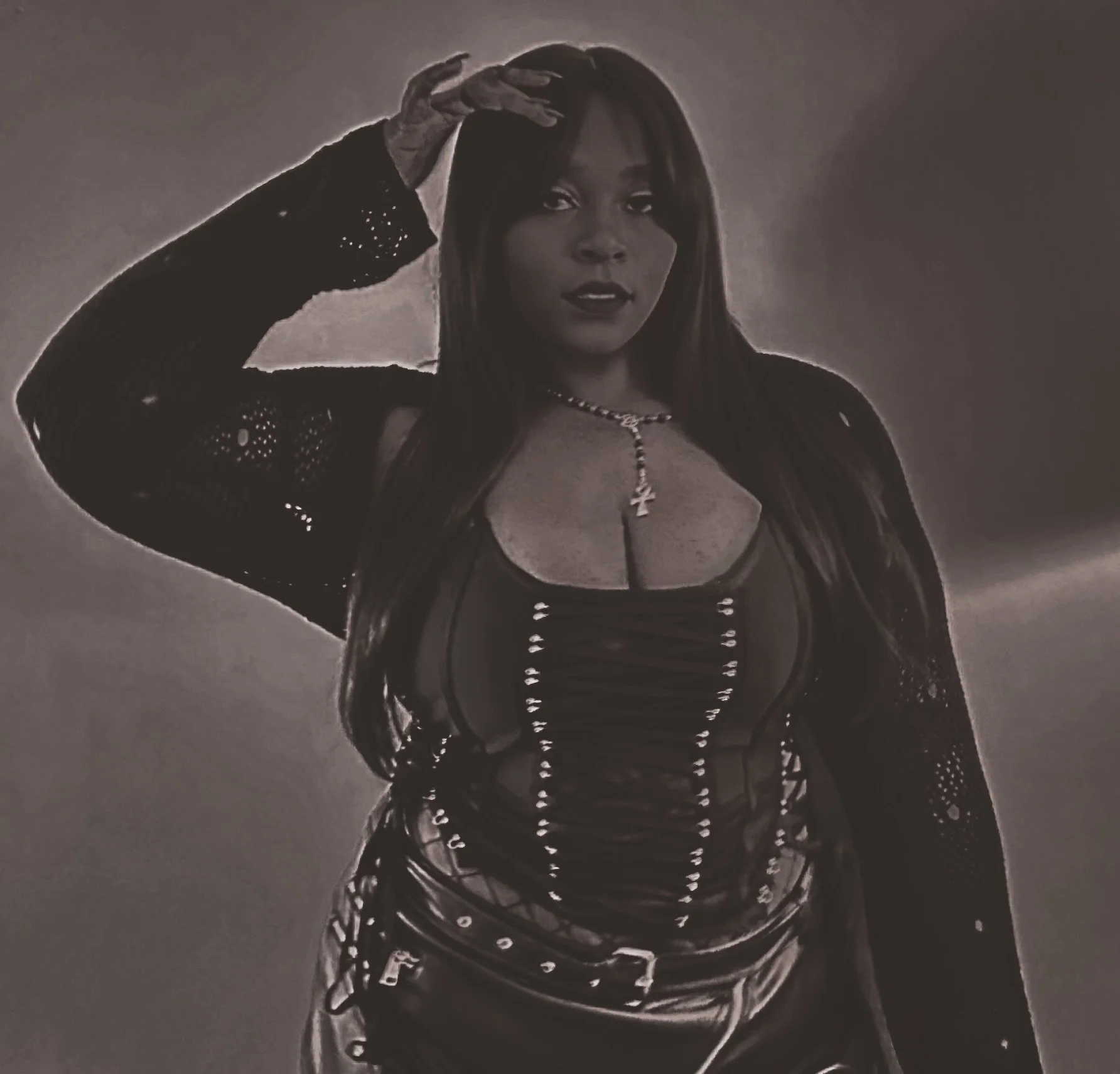A woman with long hair and light skin wearing a black lace dress and cross necklace, holding her hand above her head, looking directly at the camera with a confident expression.