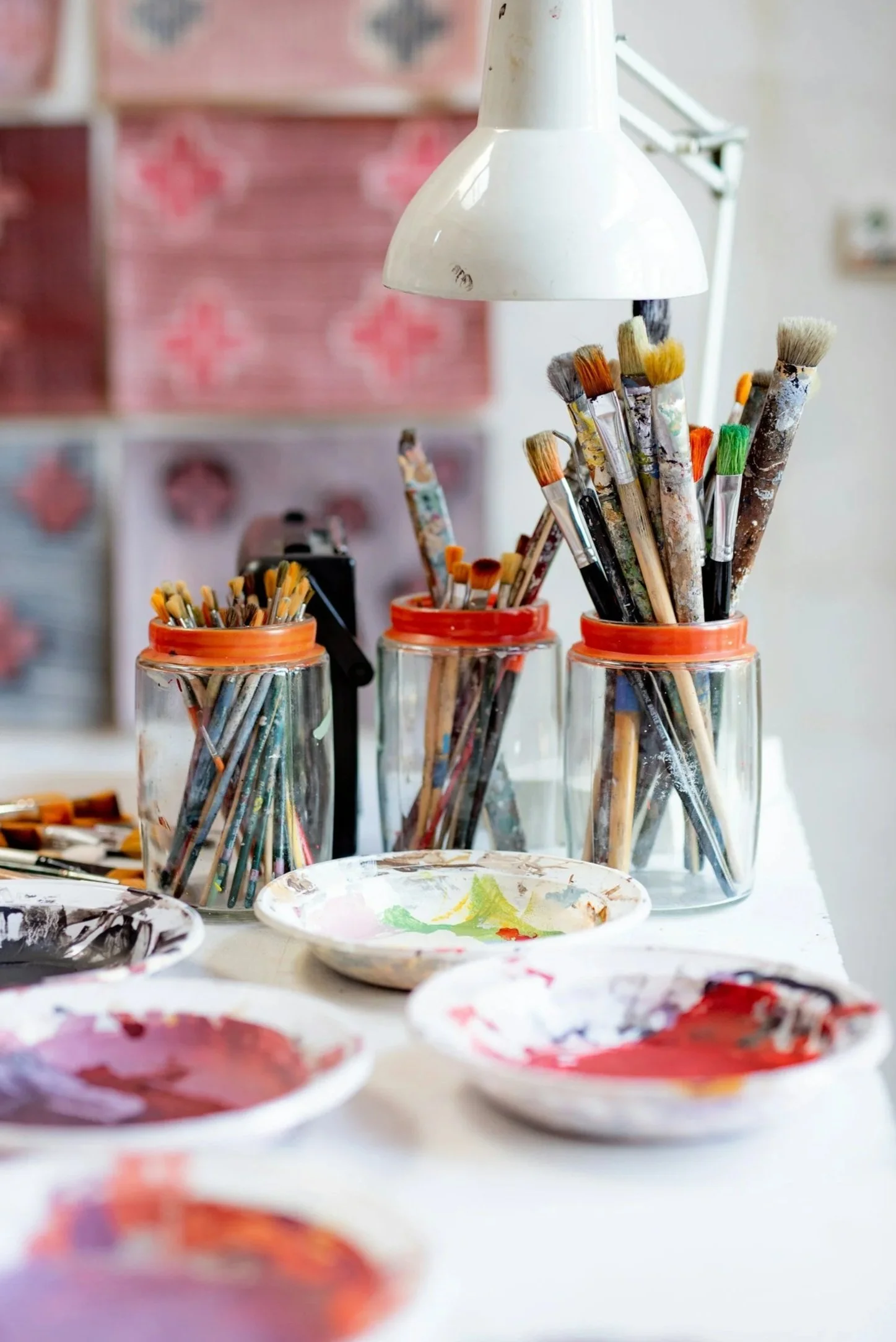 Paintbrushes in glass jars on a table with paint palettes and a white lamp overhead.