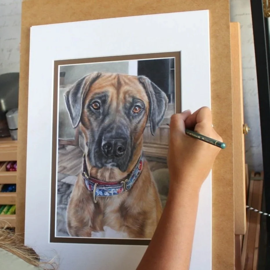An artist is painting a realistic portrait of a brown dog with a black muzzle and expressive eyes, wearing a colorful collar, on a white easel.