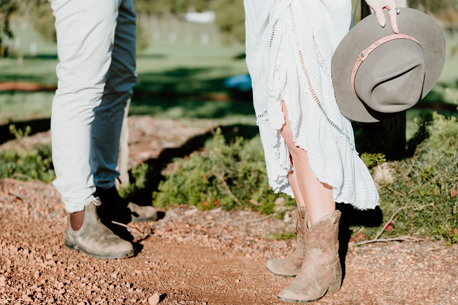 Two people standing outdoors on a dirt path. One person is wearing light-colored pants and boots, and the other person is wearing a white dress and cowboy boots, holding a gray hat.