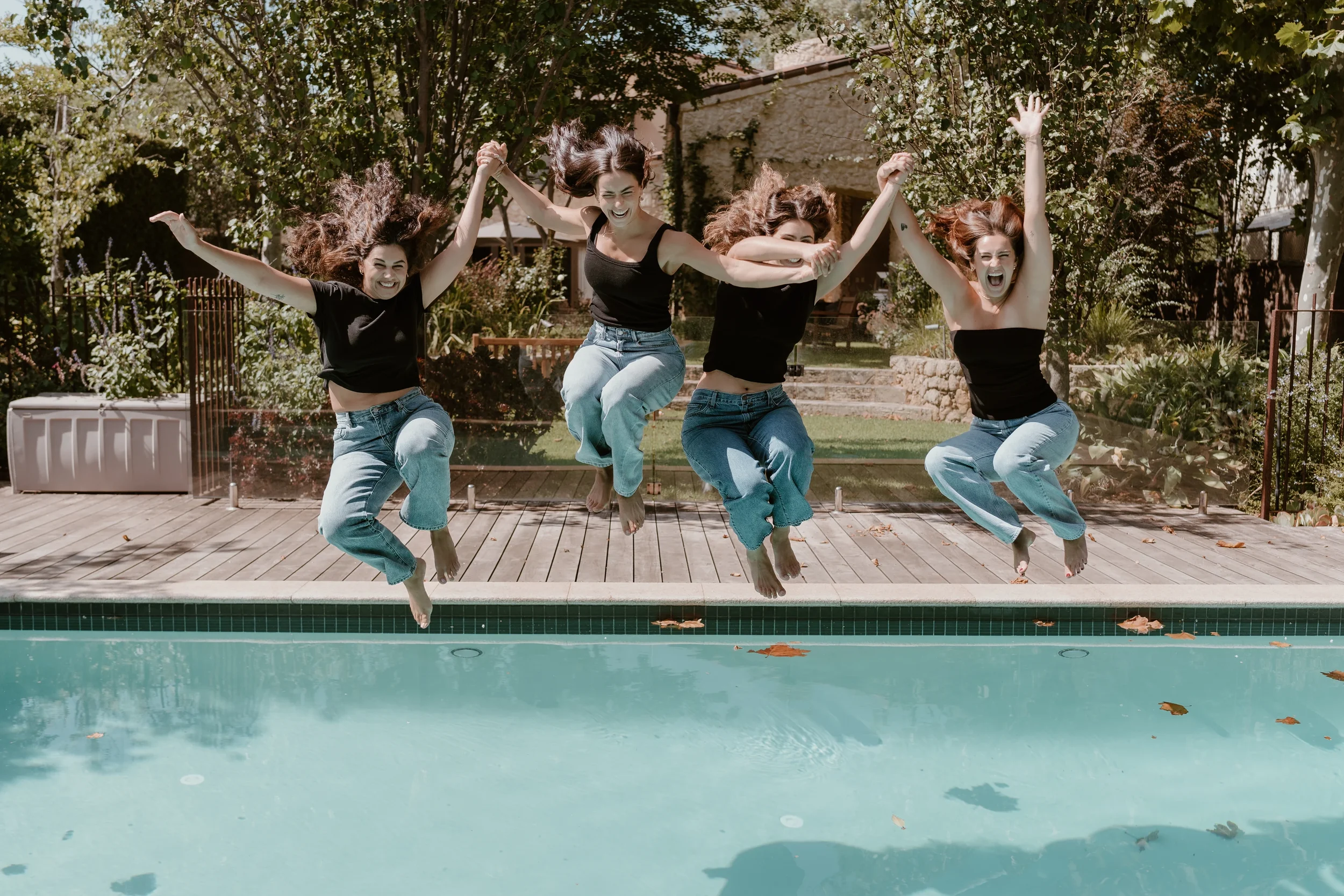 Four women jumping into a pool, smiling and holding hands, on a sunny day with trees and a garden in the background.