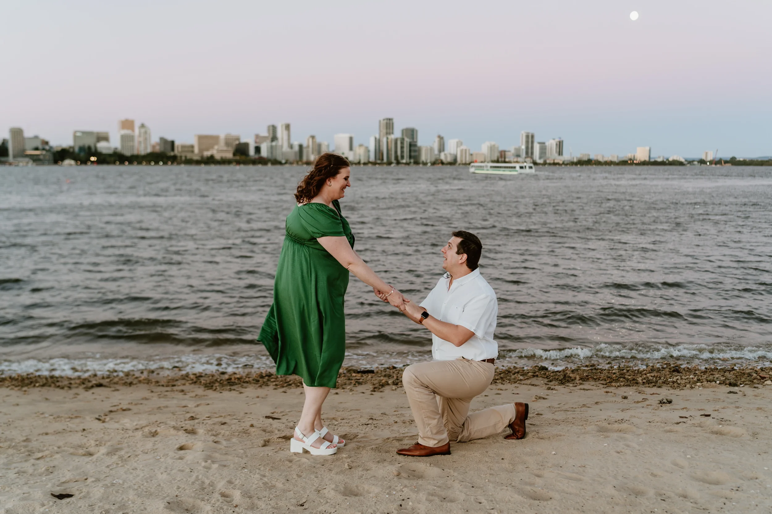 A man is proposing marriage to a woman on a beach with a city skyline and water in the background. The woman is standing, wearing a green dress and white sandals, while the man, kneeling on one knee, is holding her hands and wearing a white shirt and