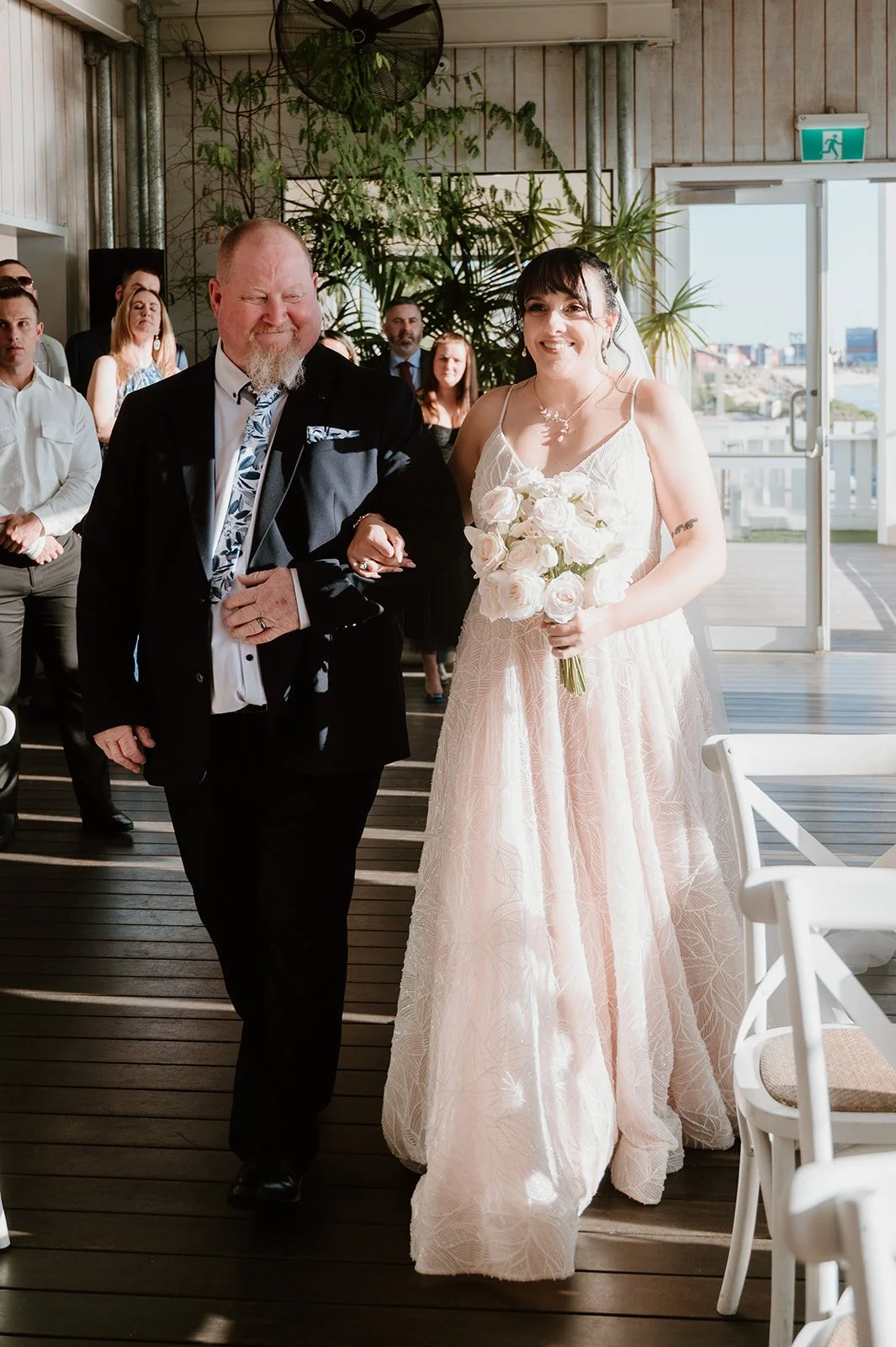 A bride in a light pink wedding dress holding a bouquet of white roses, walking down the aisle with a man in a black suit, during a wedding ceremony with guests in the background.